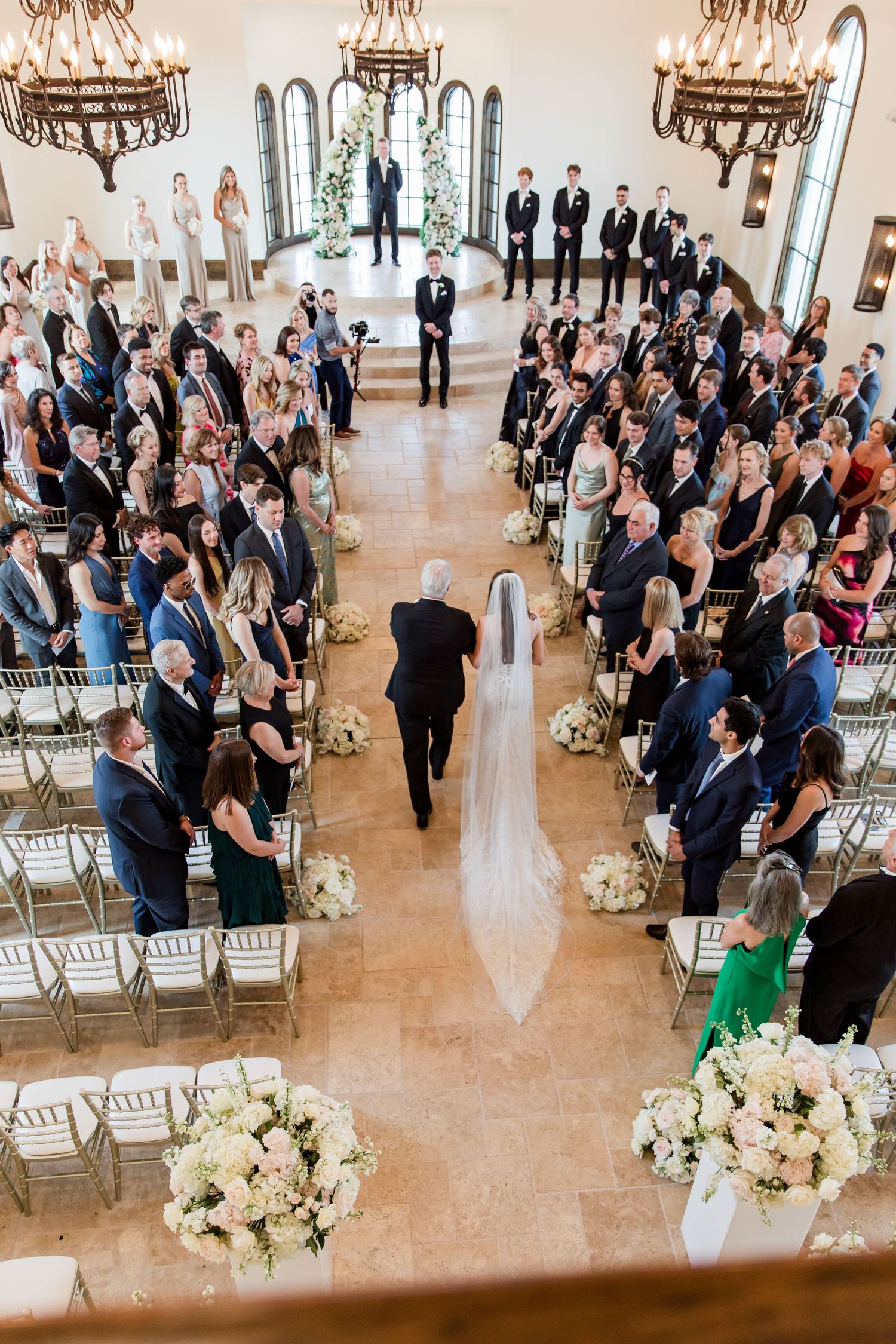 Aerial view of wedding ceremony with bride walking down aisle in chapel filled with guests and white floral arrangements.