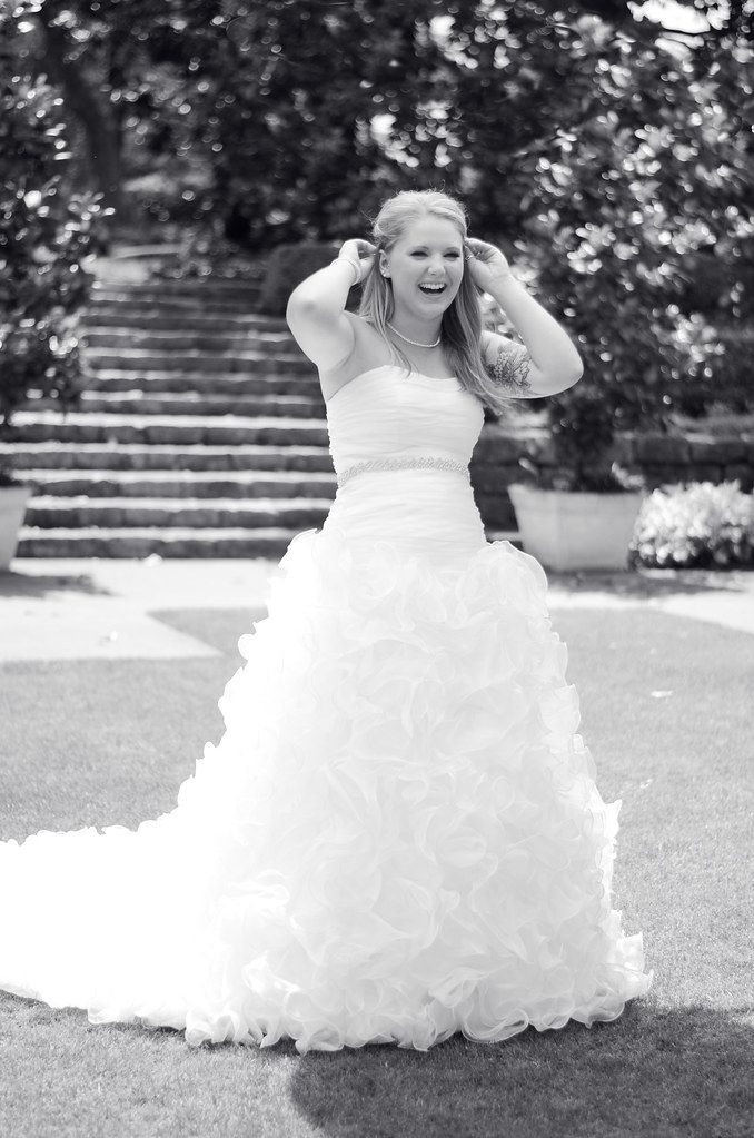 Smiling bride in a ruffled white ball gown adjusting her hair outdoors in a black and white wedding photo.