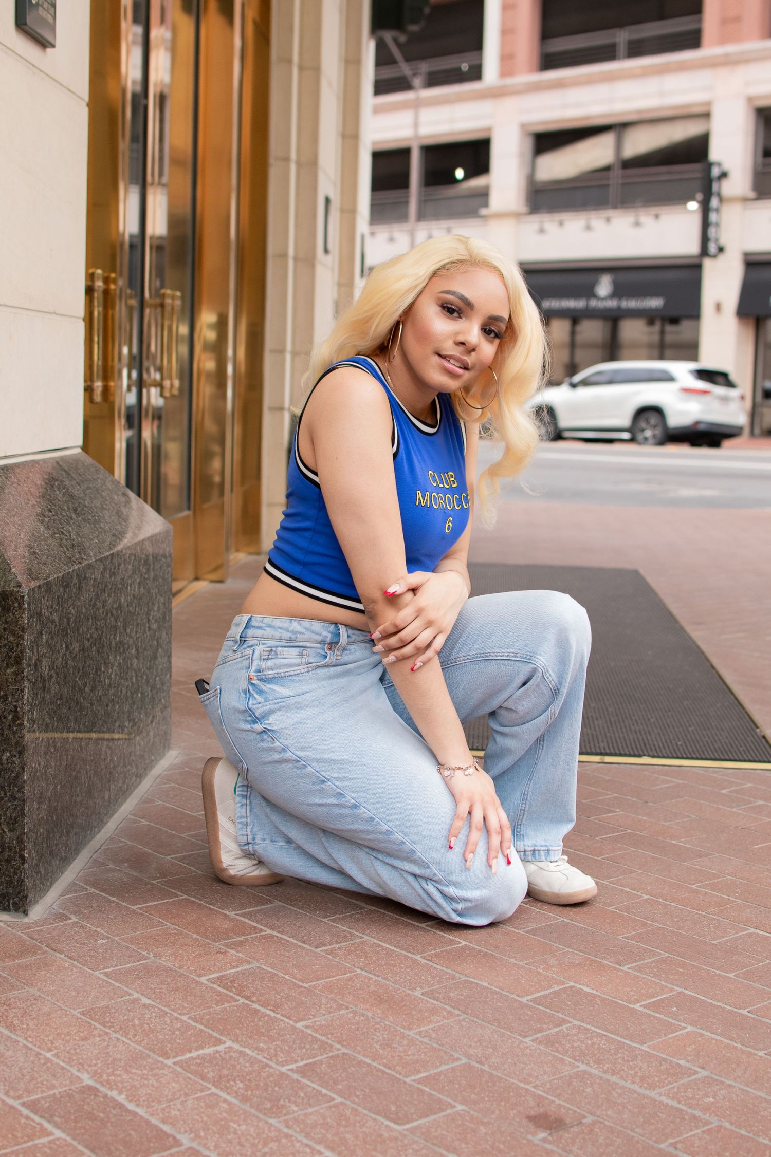 Blonde woman in blue crop top and baggy jeans crouches on urban sidewalk outside city building.