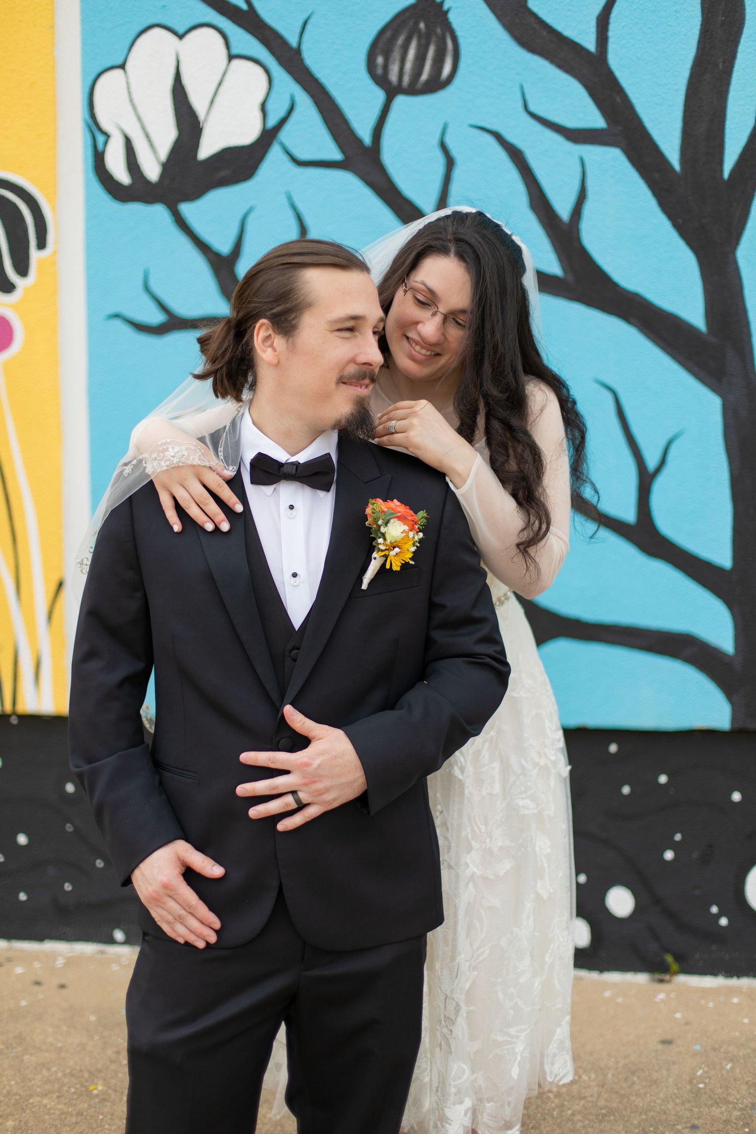 A couple embraces in front of a colorful mural featuring cotton plants and geometric designs.