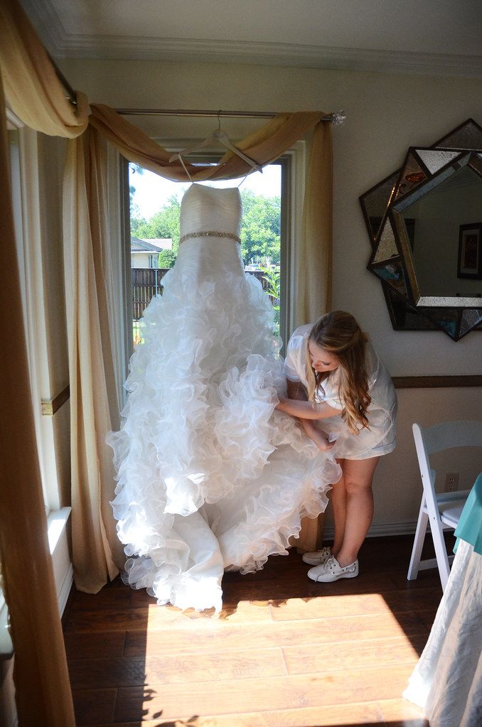 Young woman in white shirt arranging ruffled white wedding gown hanging by sunlit window.