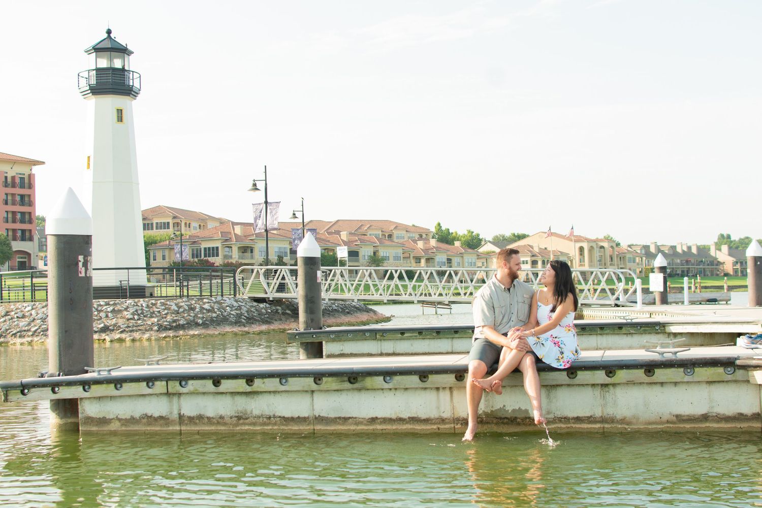 Smiling couple sitting on a dock with feet dangling near a lighthouse and marina waterfront.