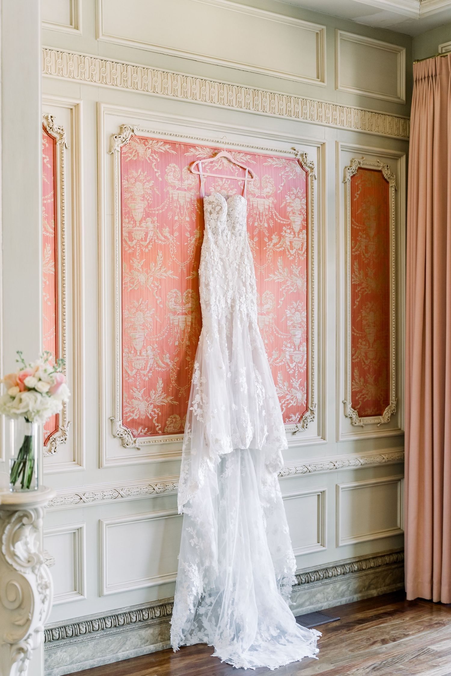 Lace wedding gown hanging on a pink velvet hanger against an ornate pink and cream paneled wall.