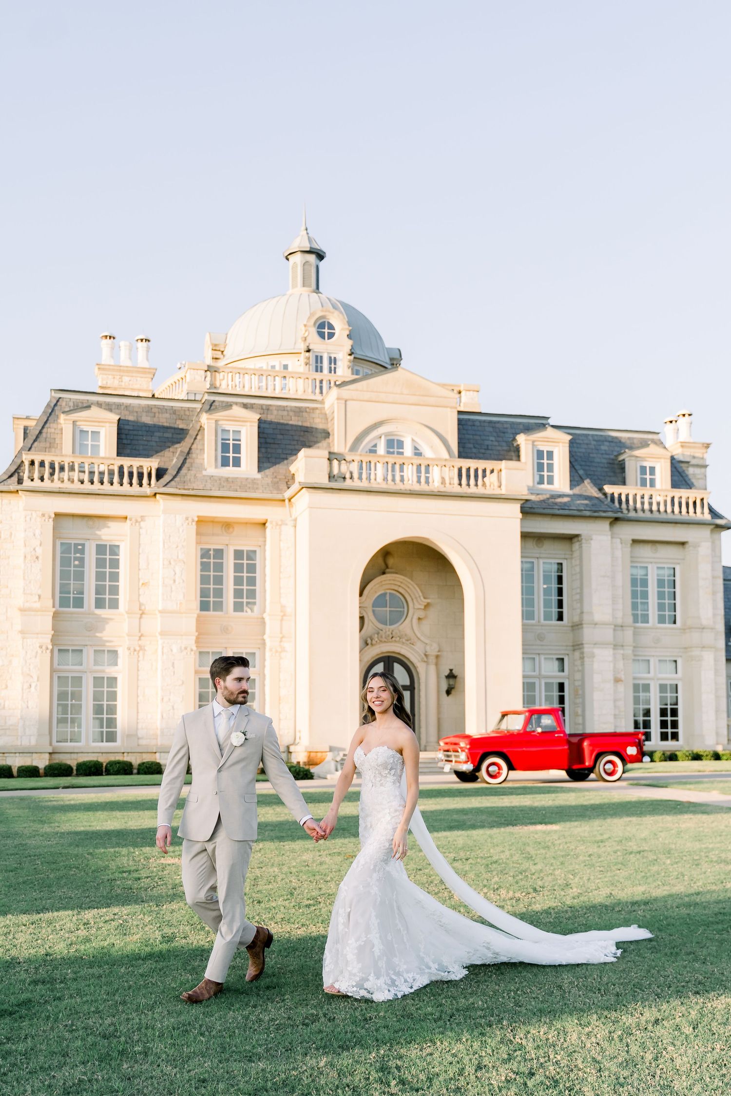 Bride and groom walking hand in hand before a grand French chateau with a vintage red truck.