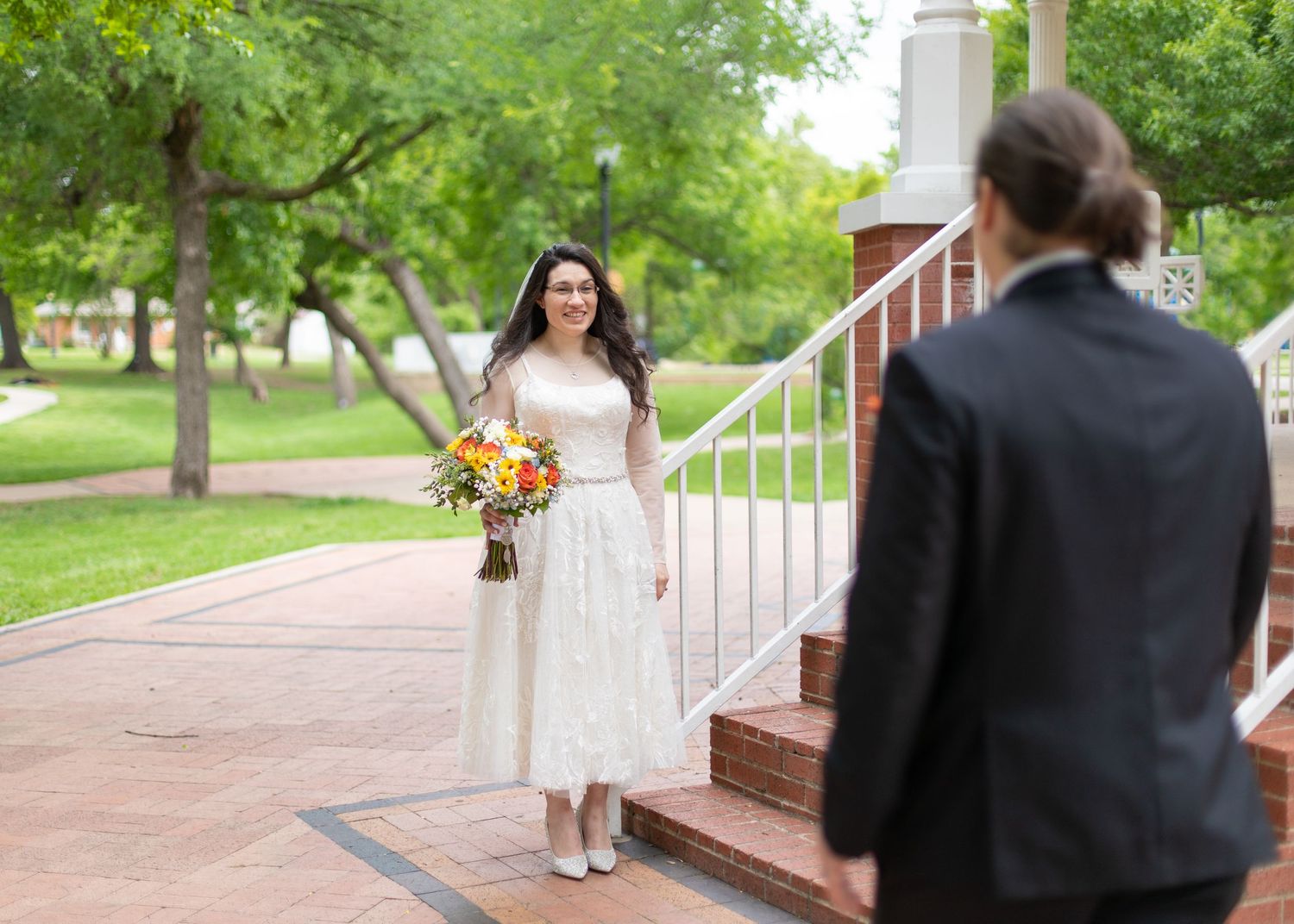 A bride in a white tea-length dress greets someone on brick steps surrounded by greenery.