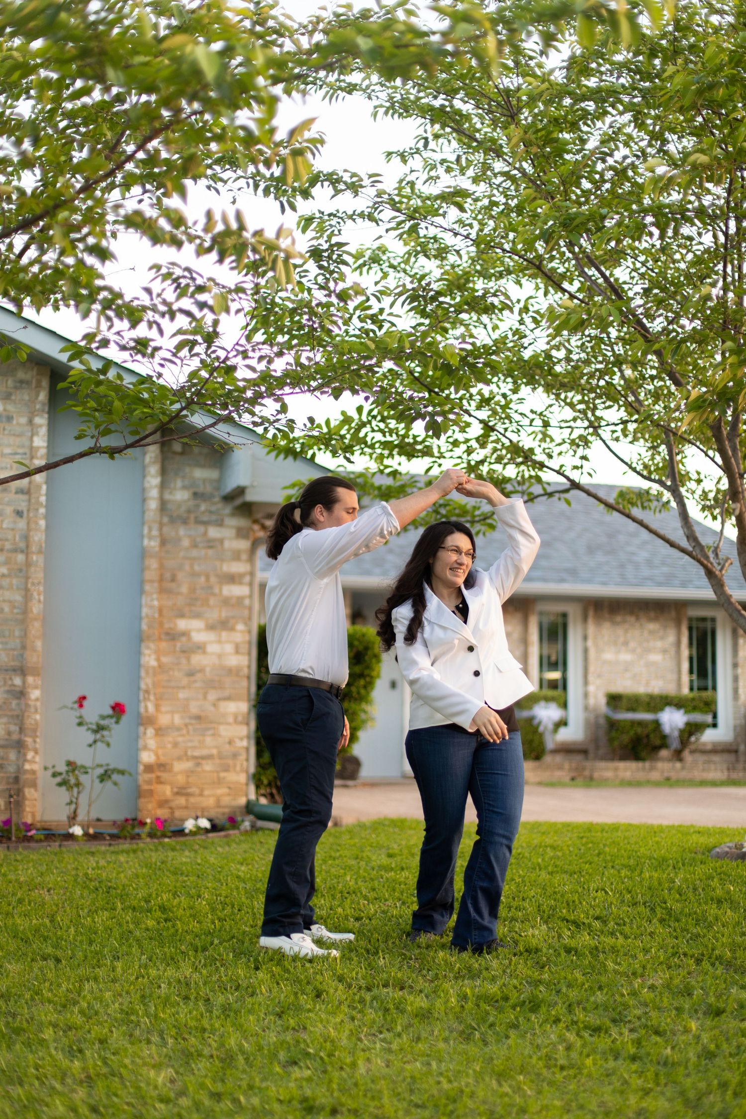 Dancing couple in matching white shirts twirling under a leafy tree in front of brick house.