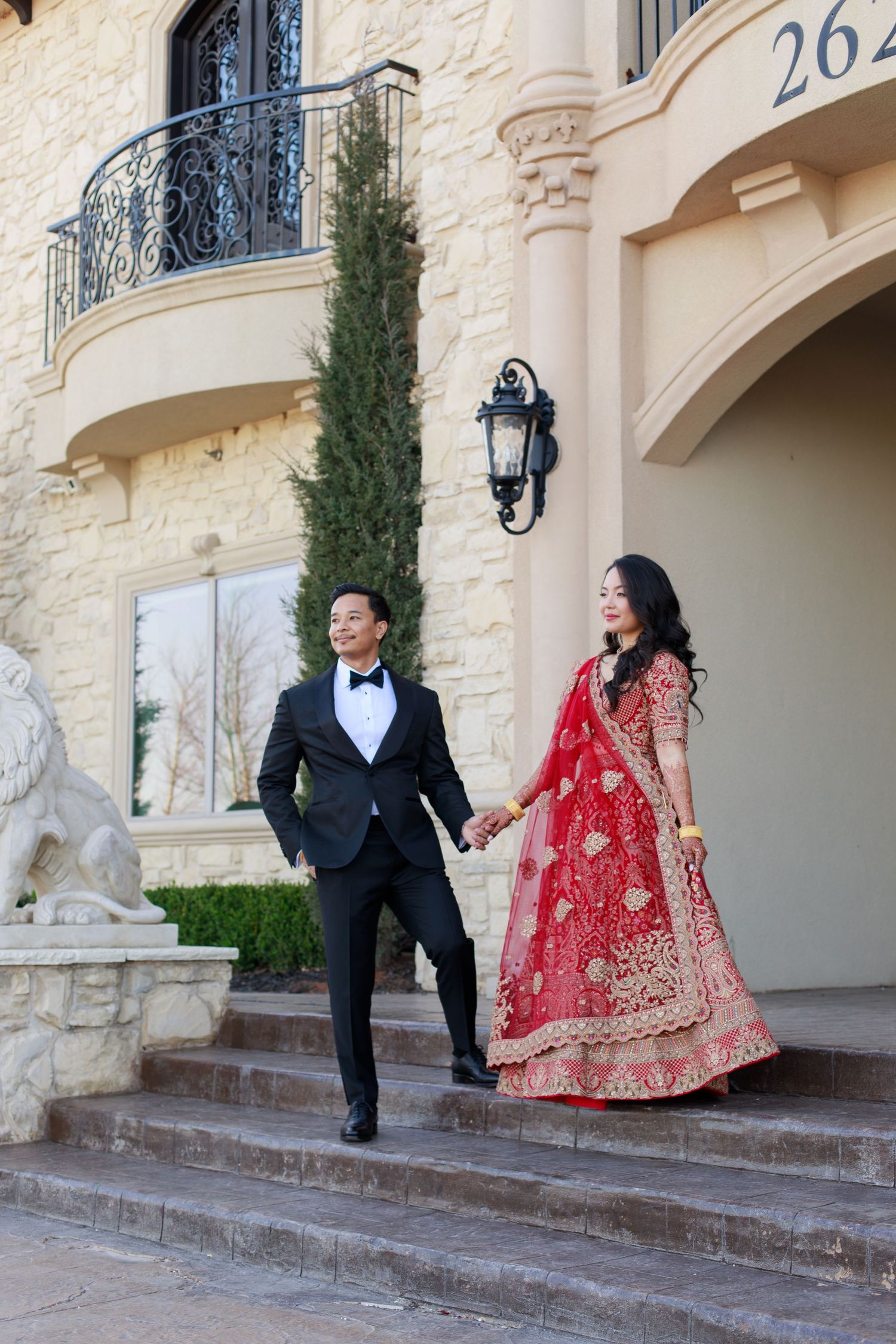 Indian couple in traditional wedding attire walk down steps of an elegant stone mansion holding hands.