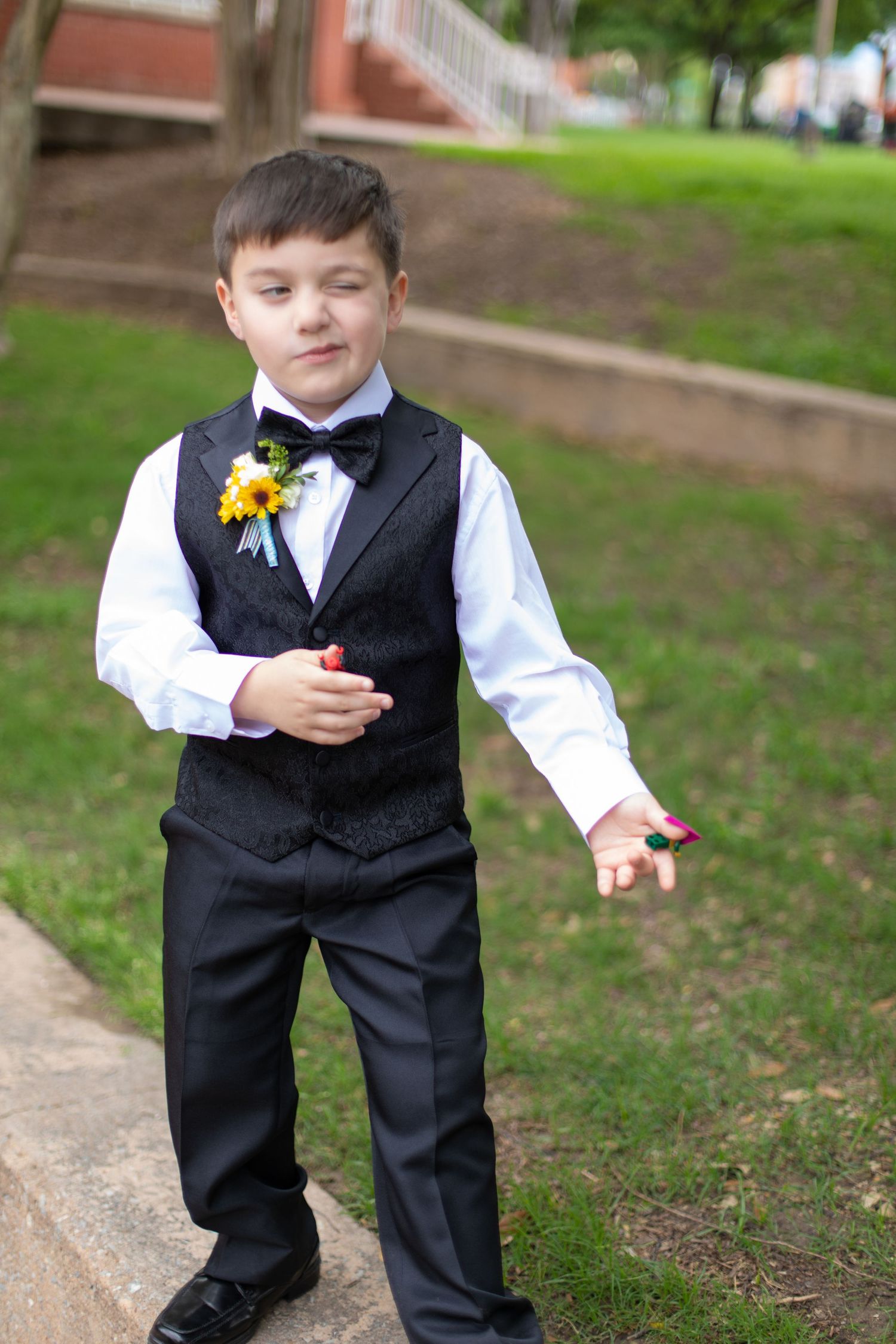 A young ring bearer in formal attire holds a yellow boutonniere while standing on brick steps outdoors.