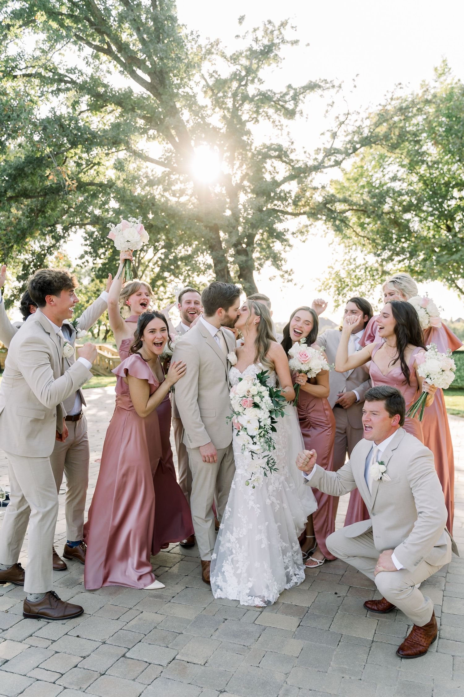 Joyful wedding party celebrating newlyweds kissing outdoors under sunlit oak trees.