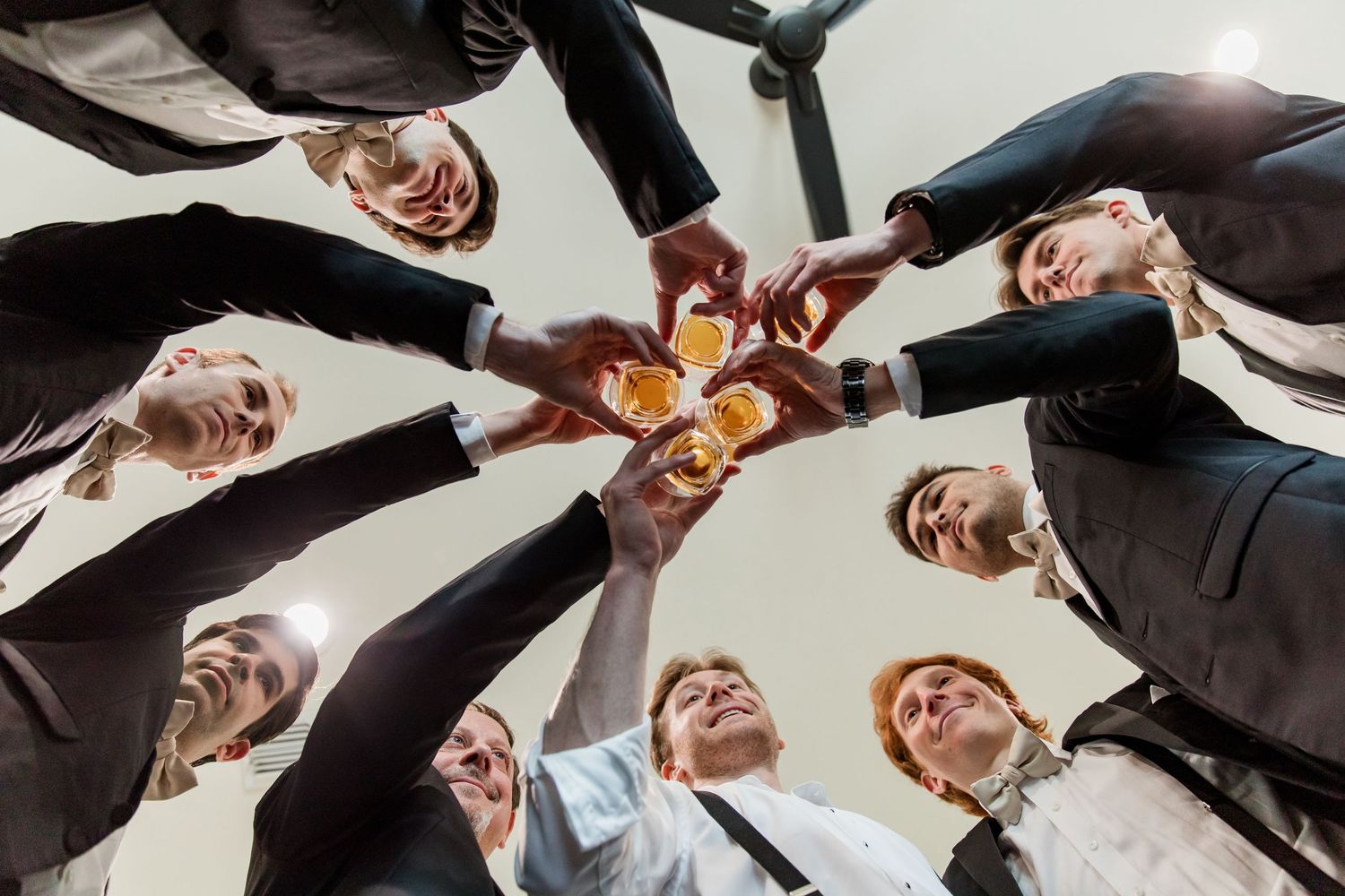 Groomsmen looking down at the camera in a circle formation while toasting their drinks. Luxury wedding photographer Gernelle Nelson, Dallas