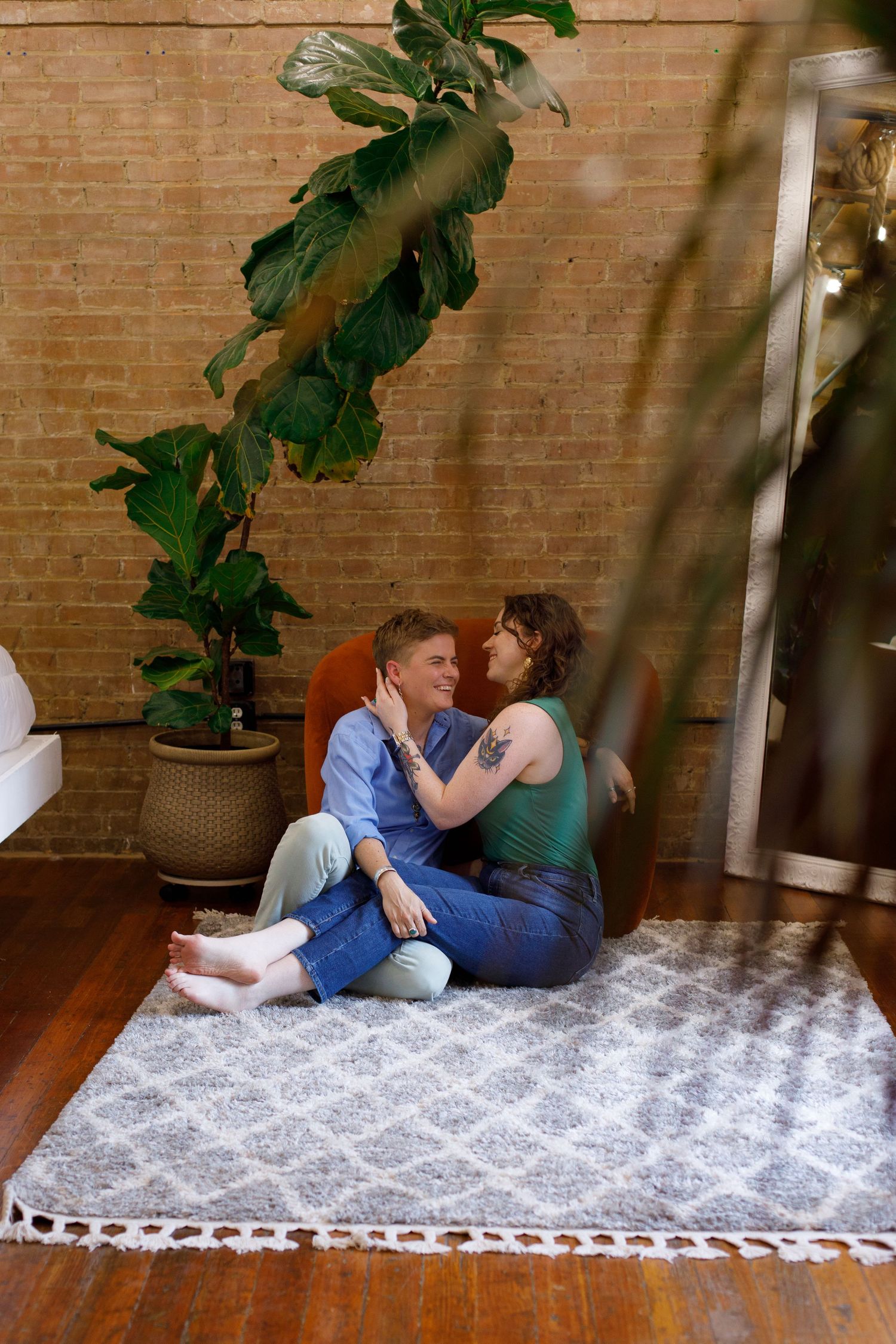Two women sitting closely together on a rug in a plant-filled brick-walled living room.