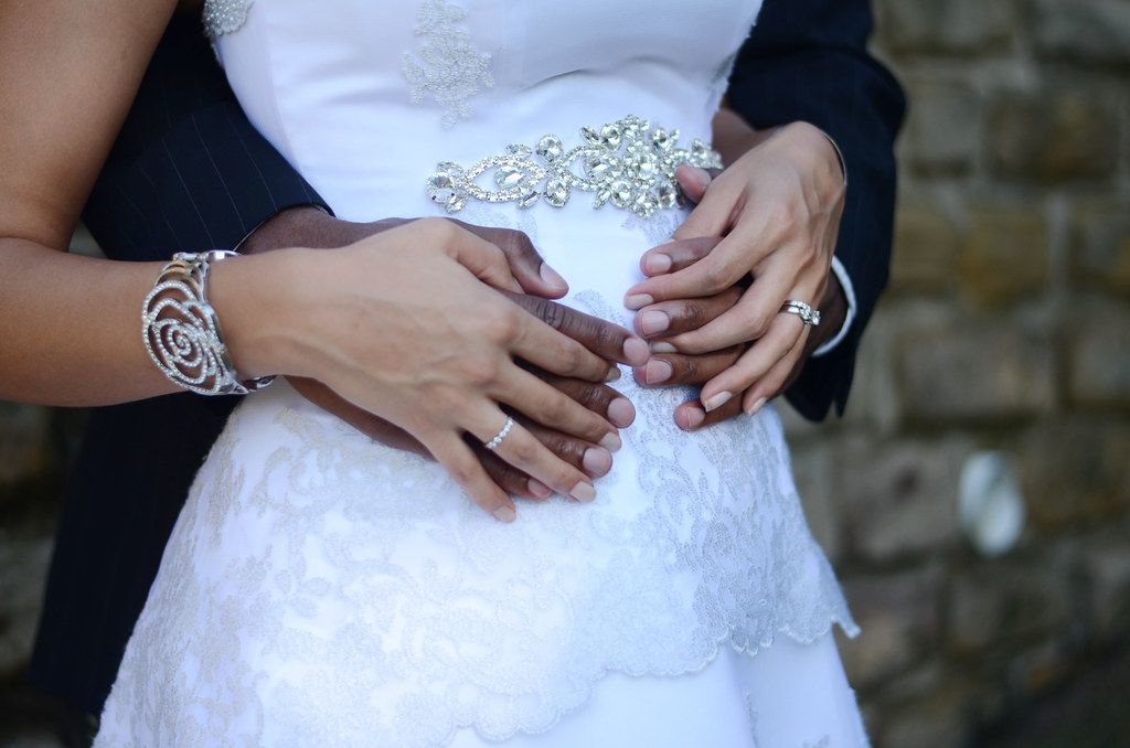 Bride and groom holding hands together on wedding dress, showcasing rings and bracelet accessories.