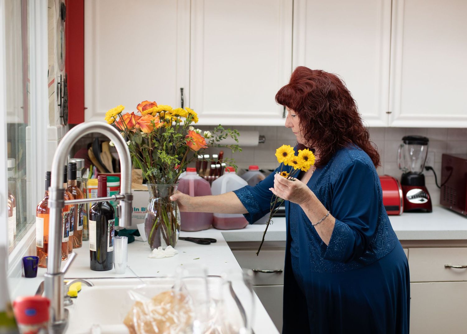Woman in blue arranging colorful flowers in a vase in a bright kitchen.