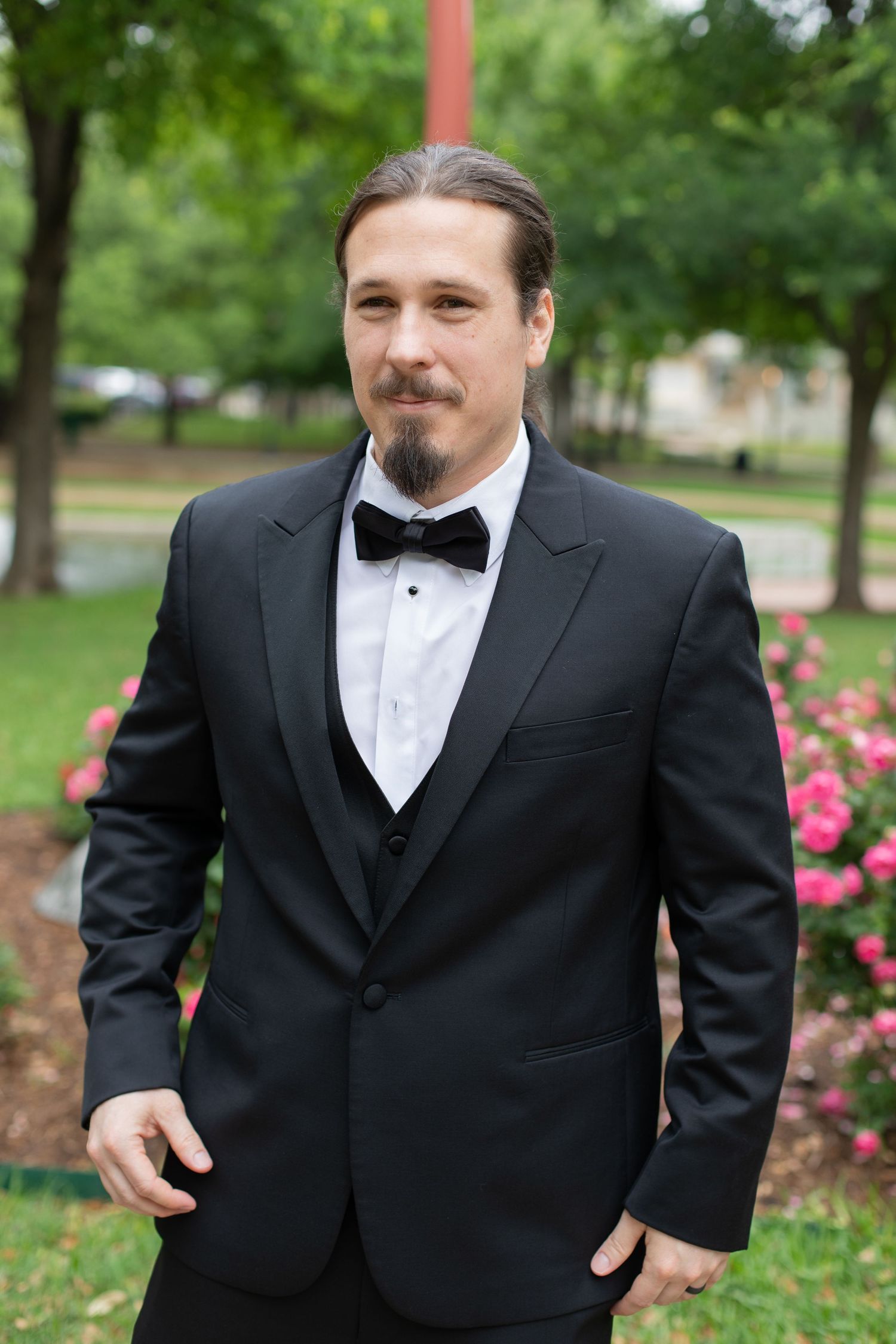 A formal portrait of someone wearing a black tuxedo with bow tie in a garden setting.