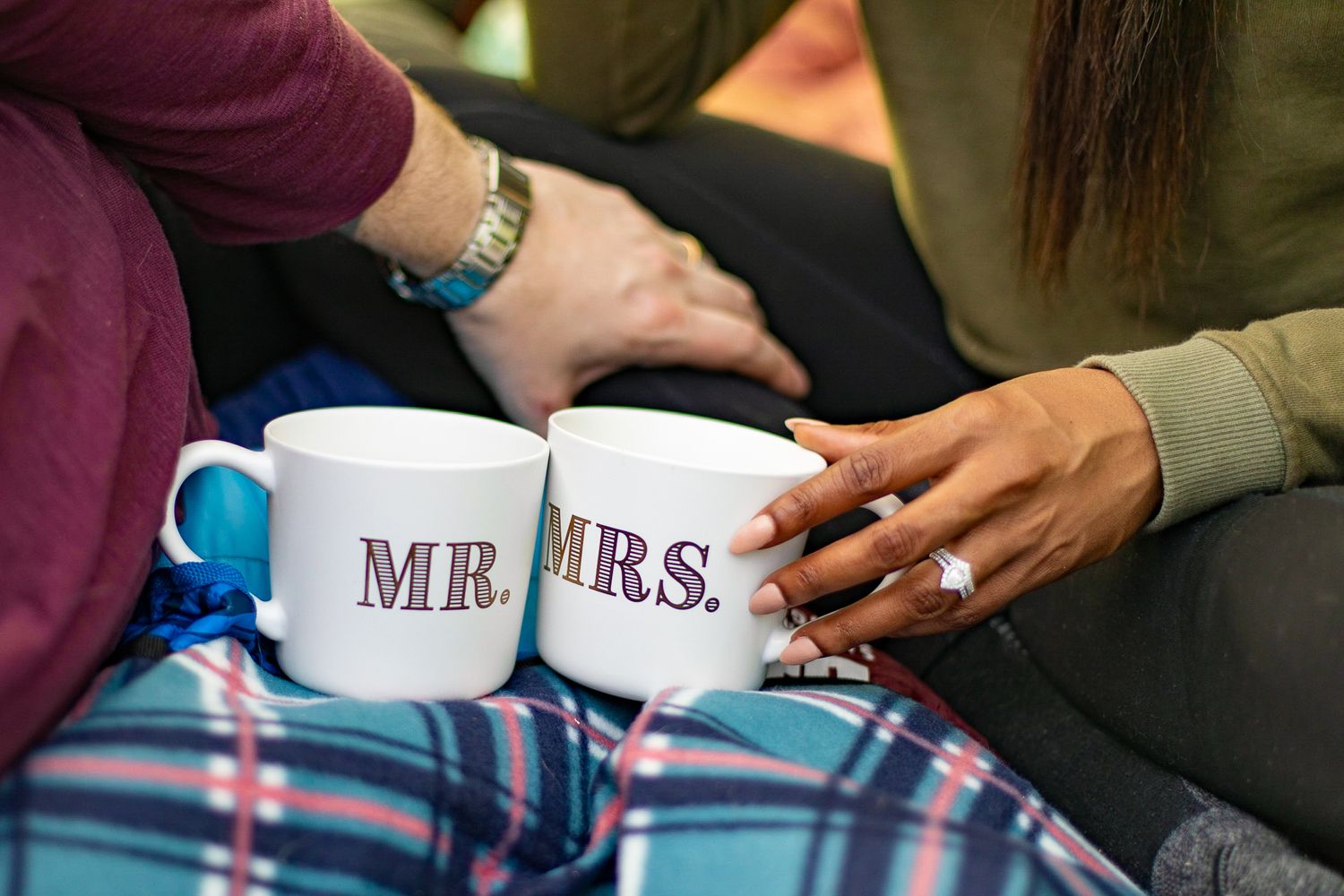 Close-up of interracial couple holding Mr. and Mrs. mugs with engagement ring visible on woman's hand.