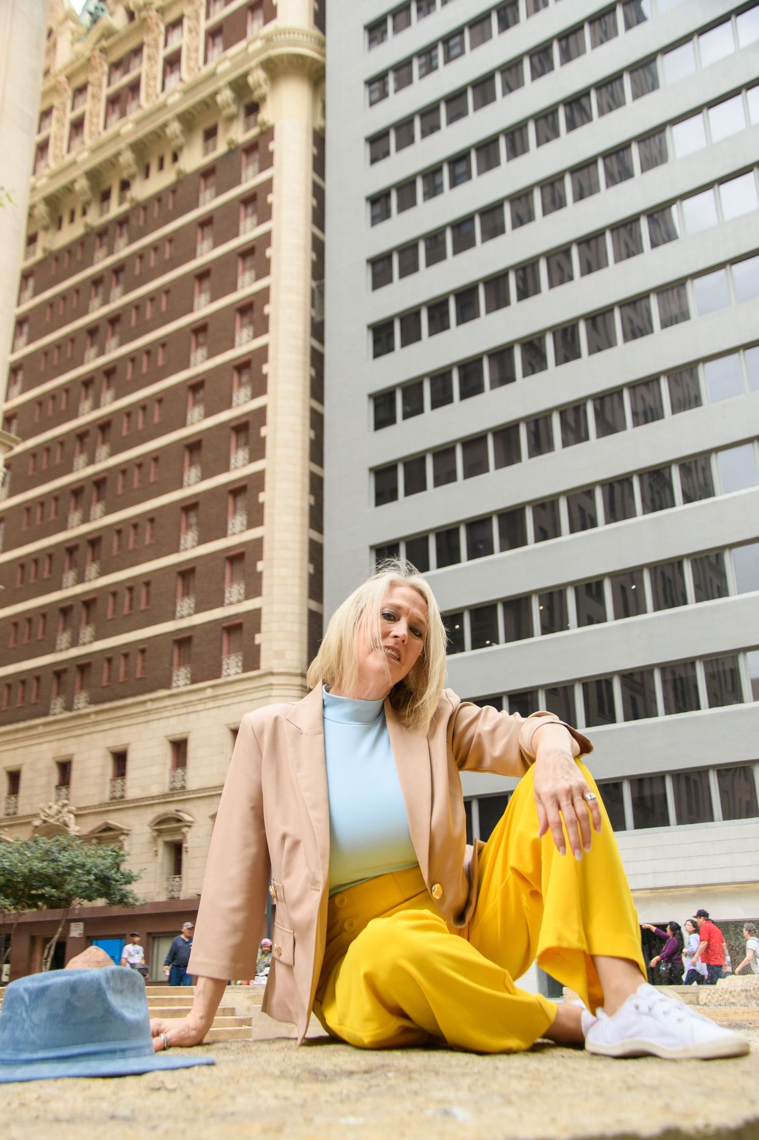 Blonde woman in yellow wide-leg pants and pink blazer poses against urban skyscraper backdrop.