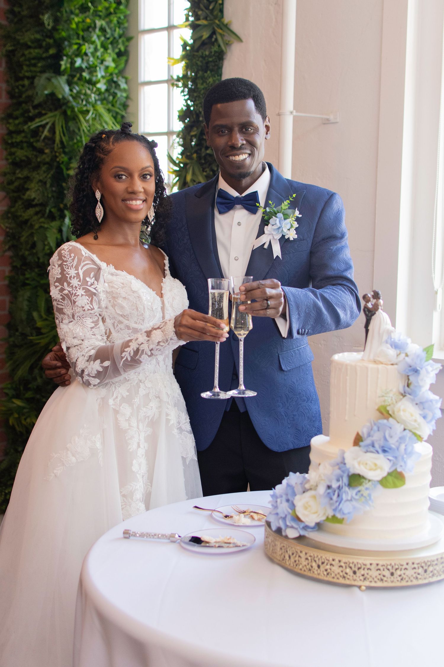Bride and groom toast with champagne flutes beside a white and blue hydrangea wedding cake.