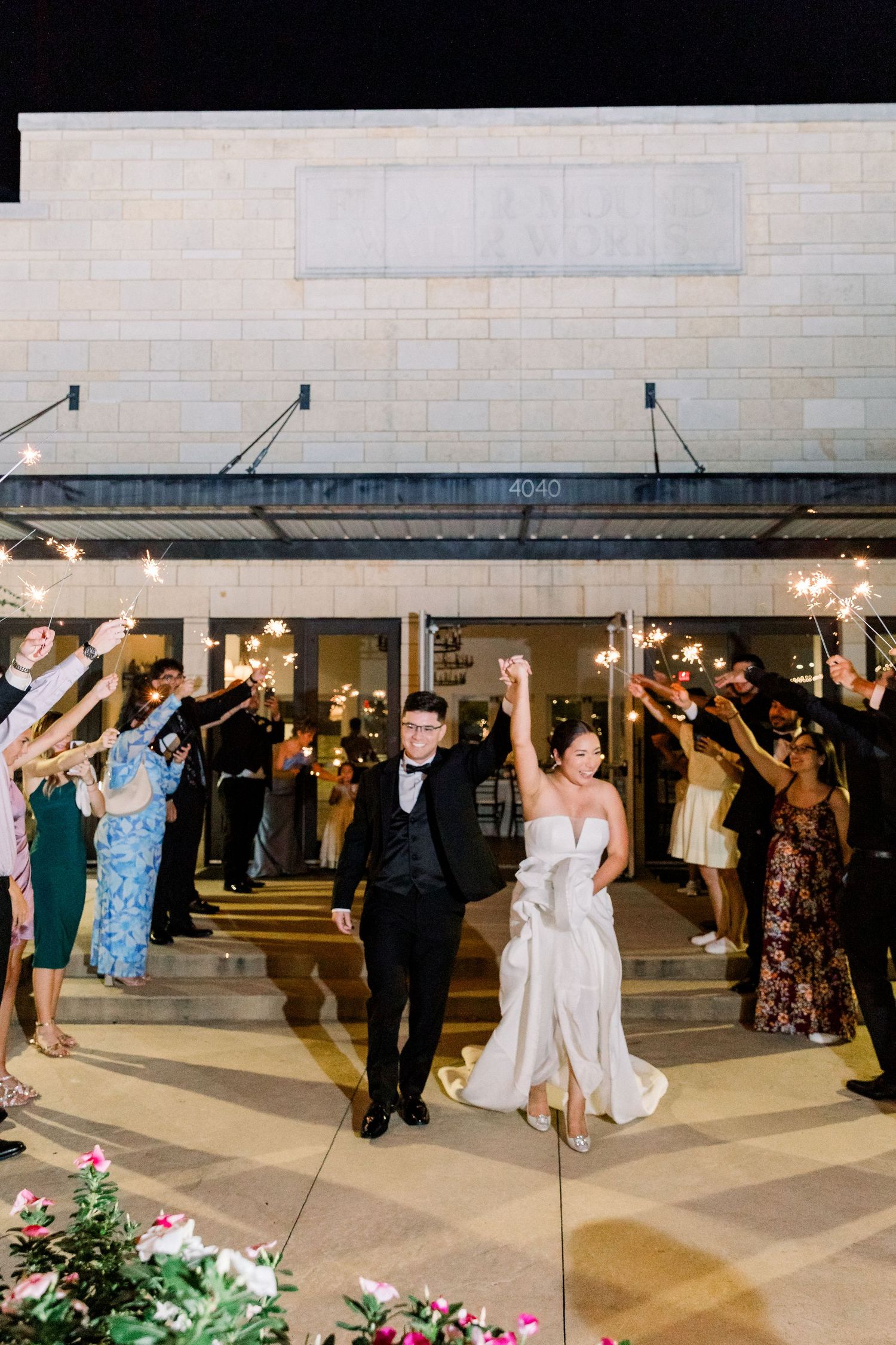 Bride and groom exit wedding venue through sparkler tunnel at night holding hands triumphantly.