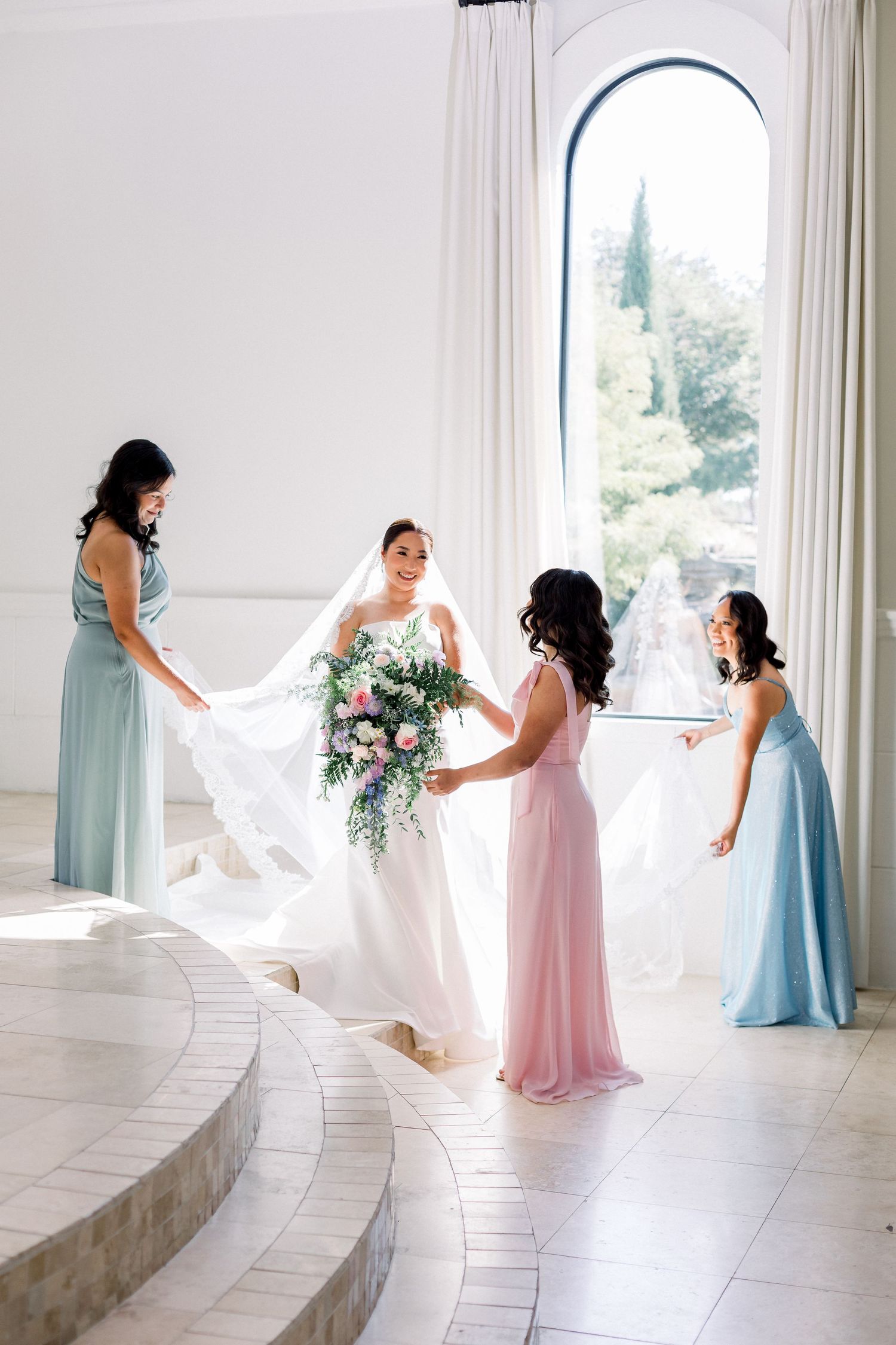 Bride in white gown holds floral bouquet surrounded by bridesmaids in blue and pink dresses in elegant white venue.