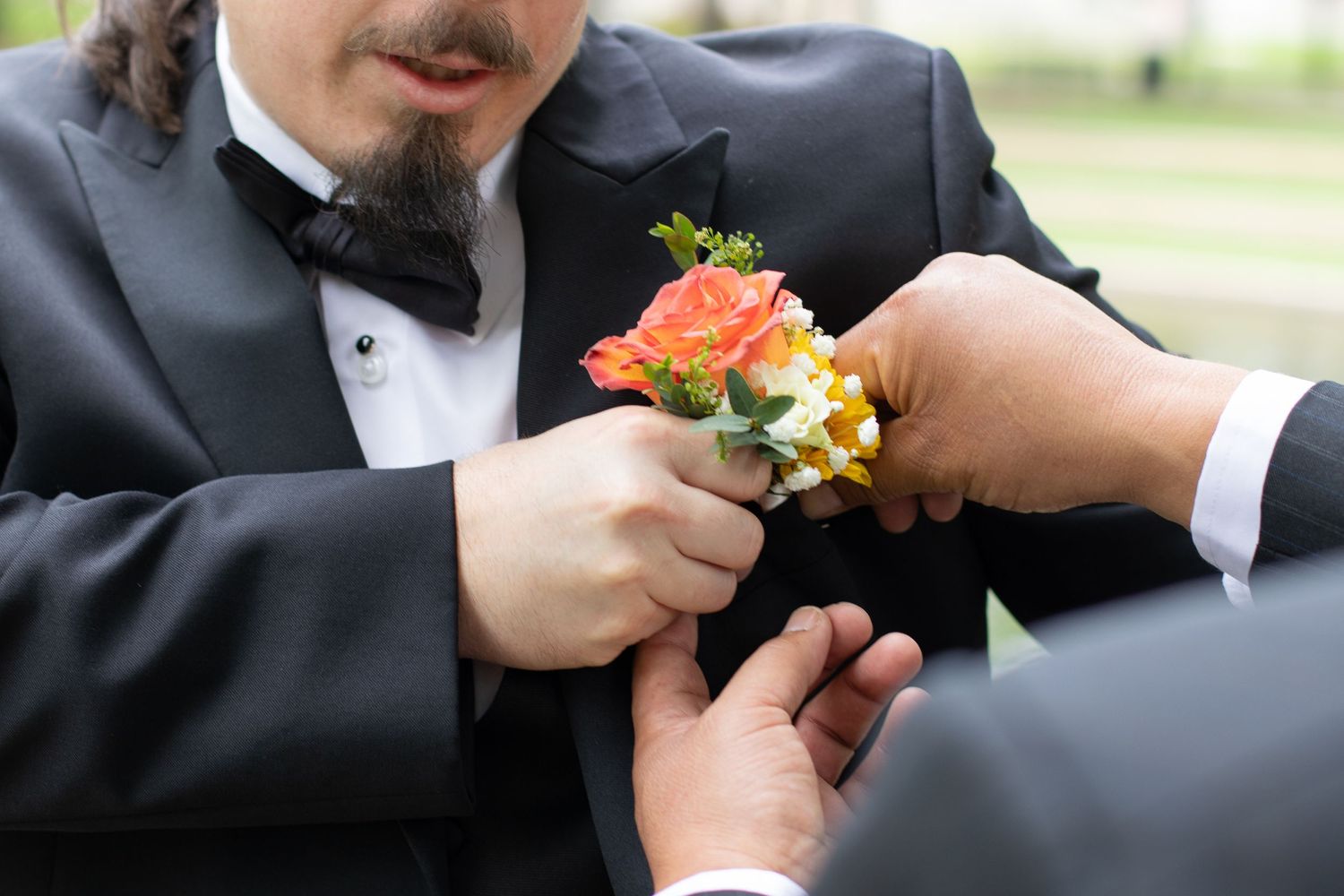 Close-up shot of someone pinning a coral and yellow boutonniere onto a formal black tuxedo.