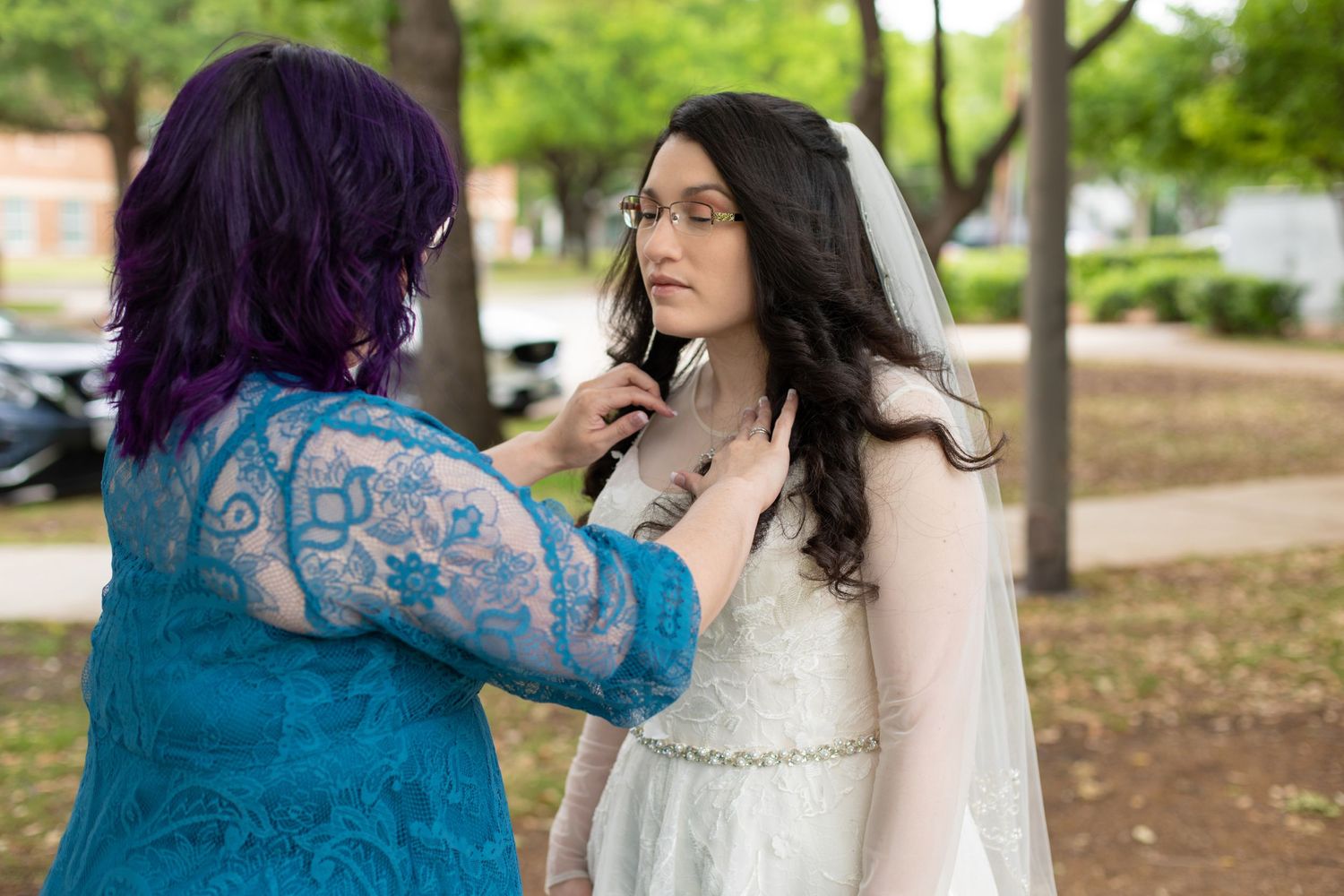 A person in a turquoise lace dress helps adjust the veil of someone wearing a white wedding gown outdoors.