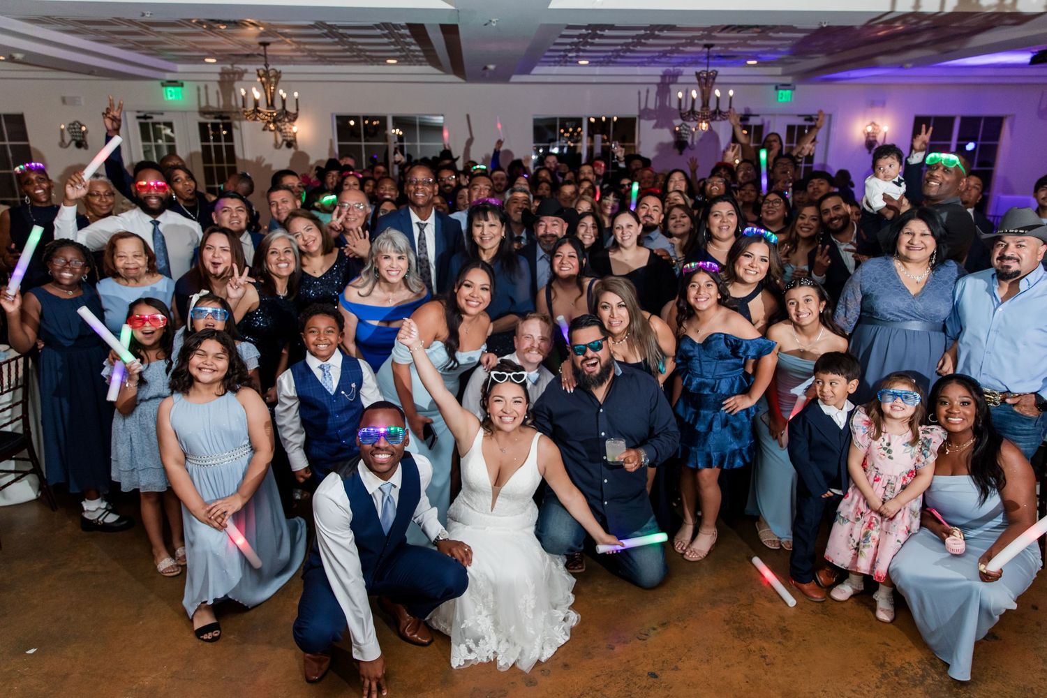 Wedding guests take a group photo to celebrate on a lively dance floor at an evening reception with purple lighting. Aristide - Flower Mound