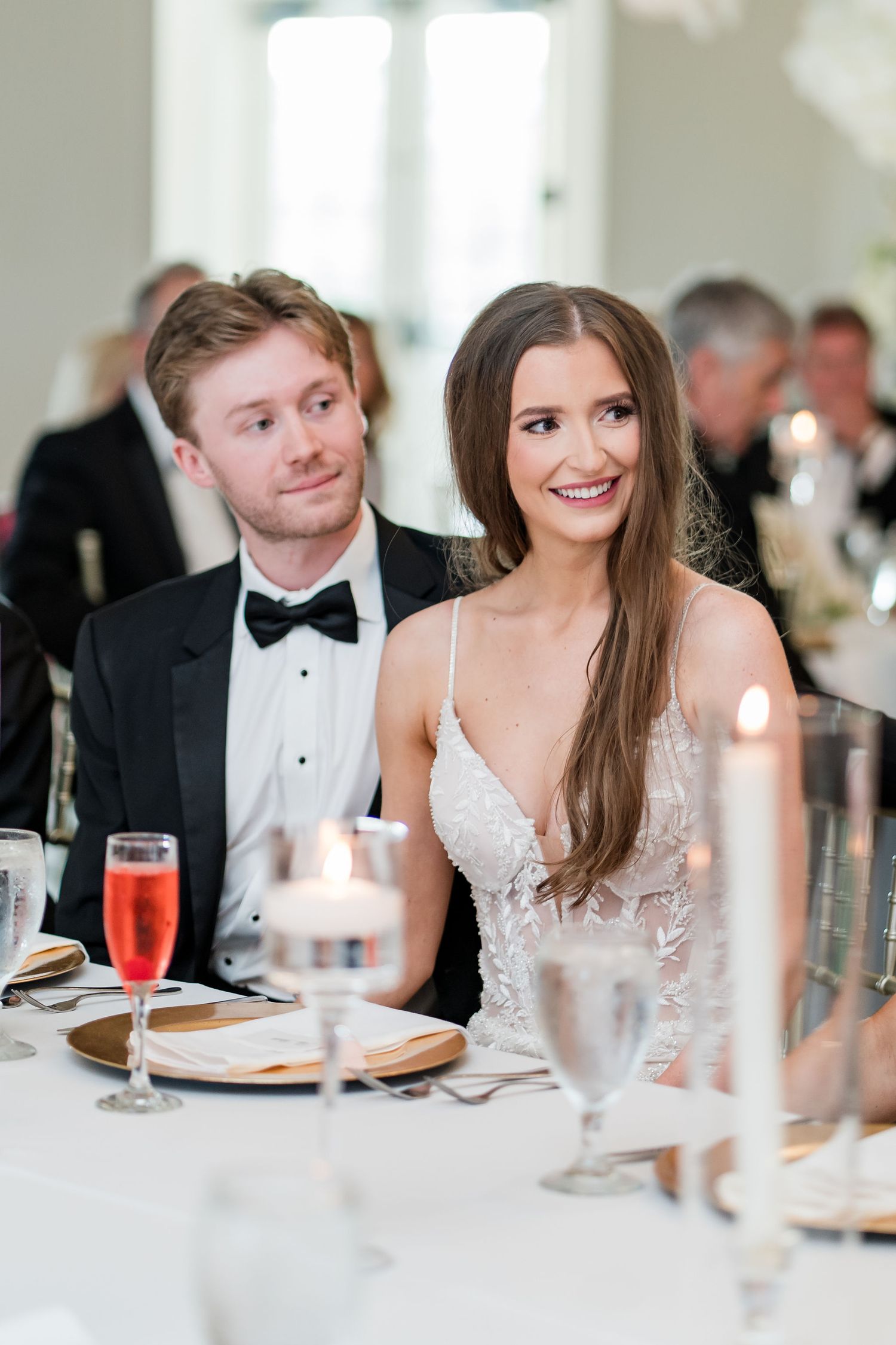 Bride and groom seated at wedding reception table with candles and champagne glasses, bride in lace gown smiling.