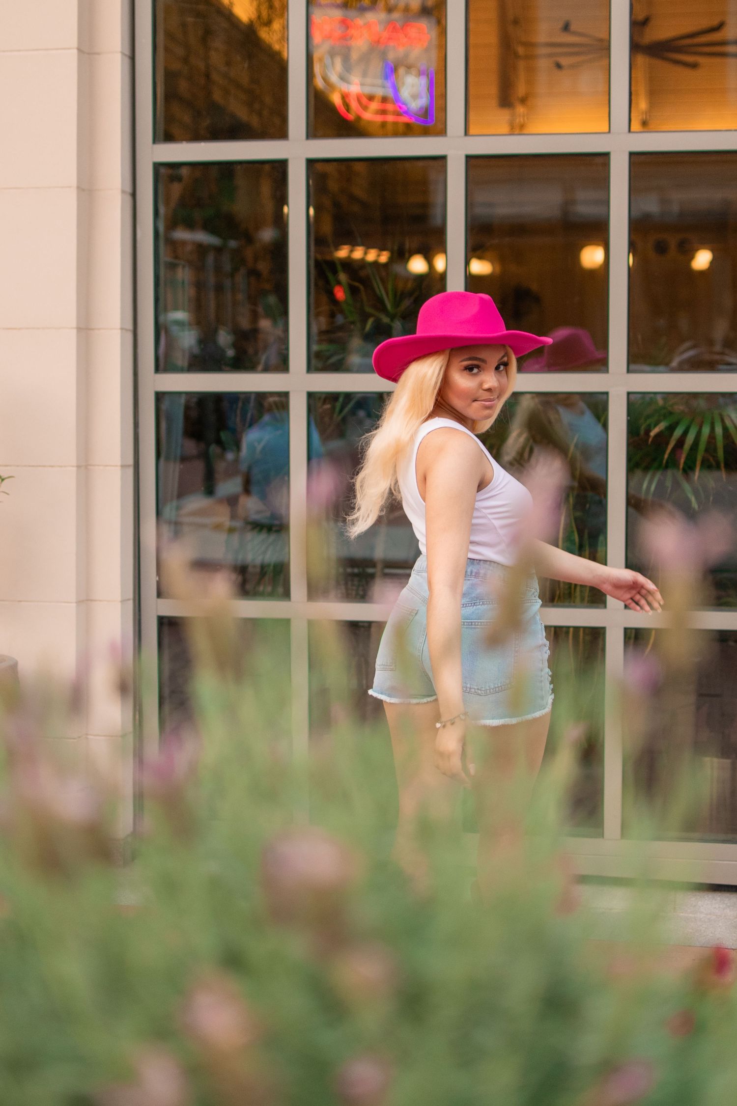 Smiling blonde woman in pink hat and white outfit poses near large window with flowers in foreground.