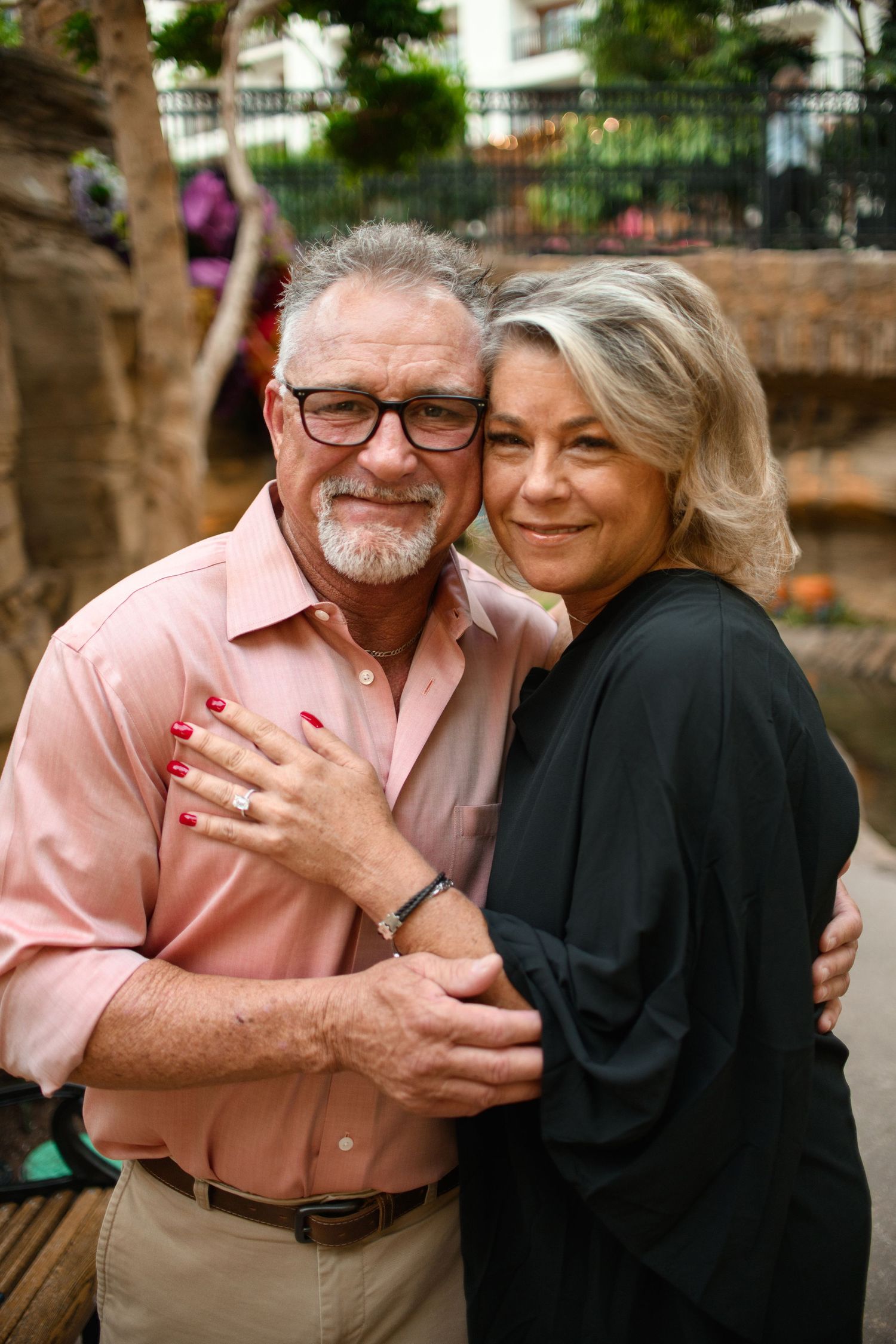 Couple in pink shirt and black top embracing outdoors against stone wall backdrop.