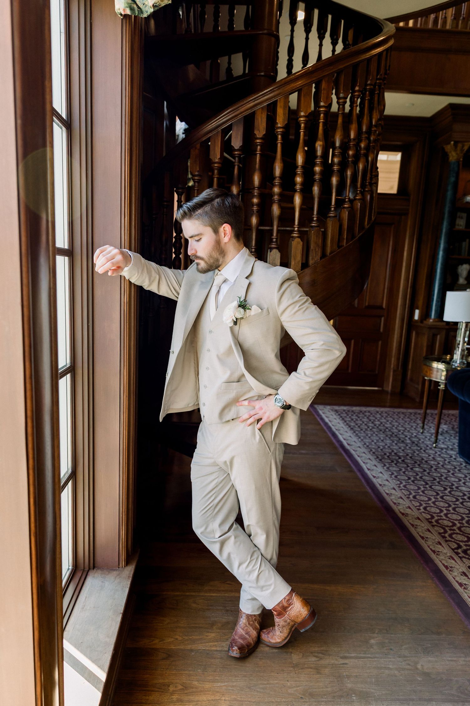 Stylish groom in tan suit leaning against a door frame inside an elegant wooden staircase foyer.