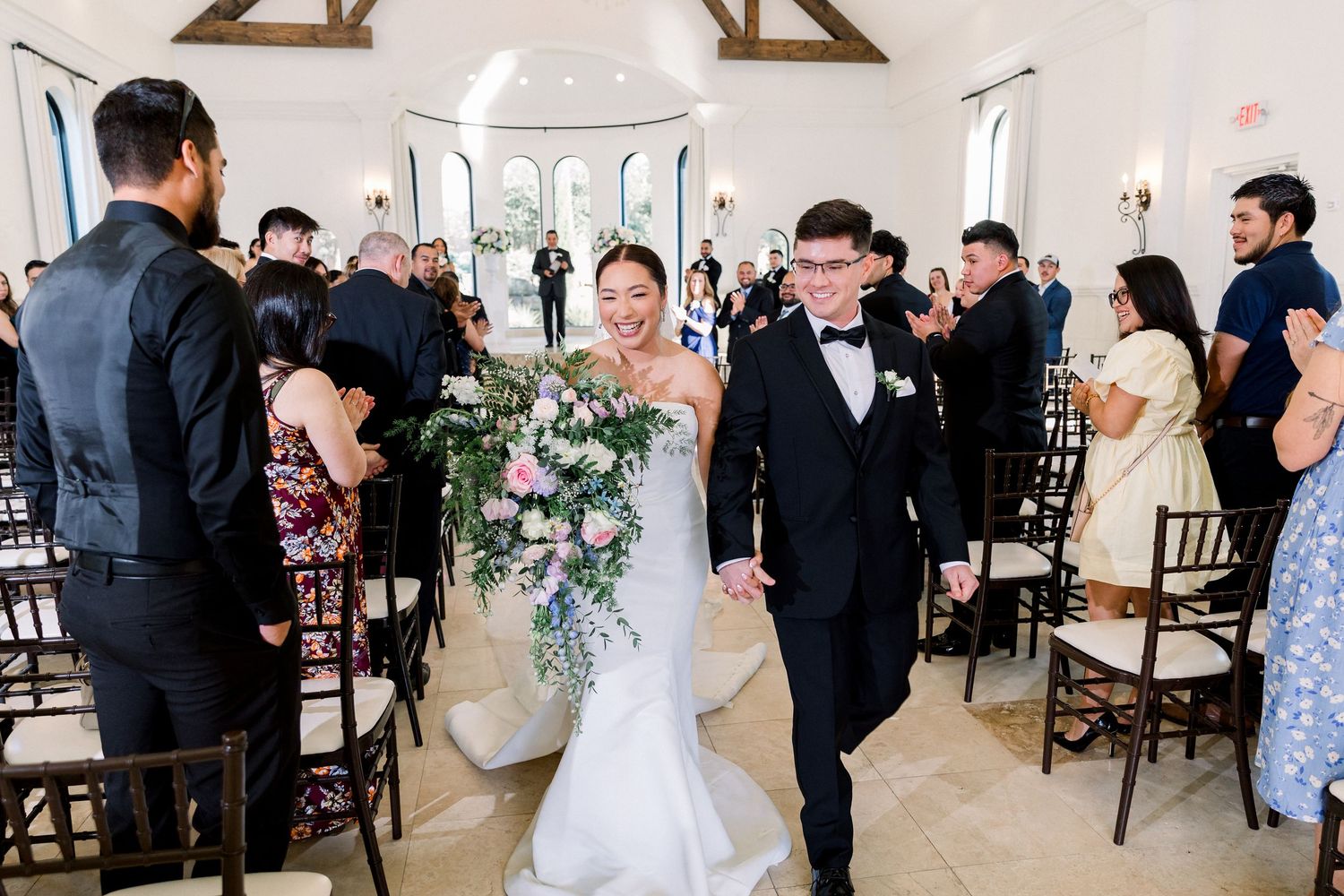 Smiling bride with lush bouquet and groom in bow tie walk joyfully down chapel aisle.