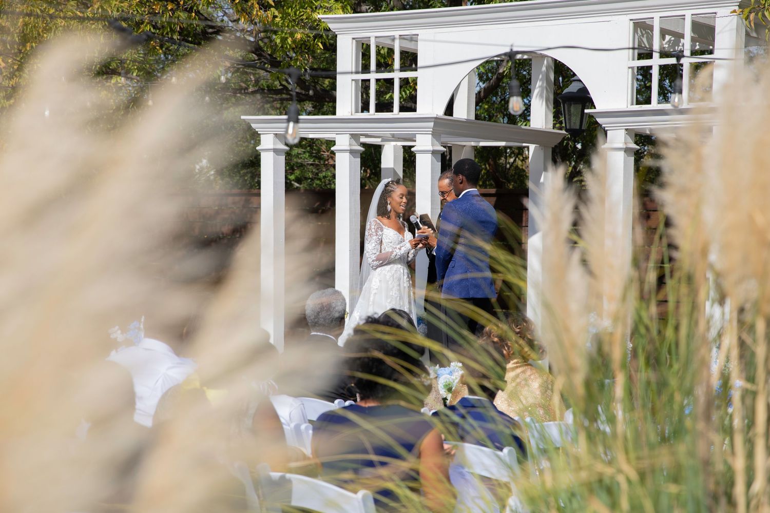 Outdoor wedding ceremony at a white pergola gazebo with guests seated, photographed through tall ornamental grasses.