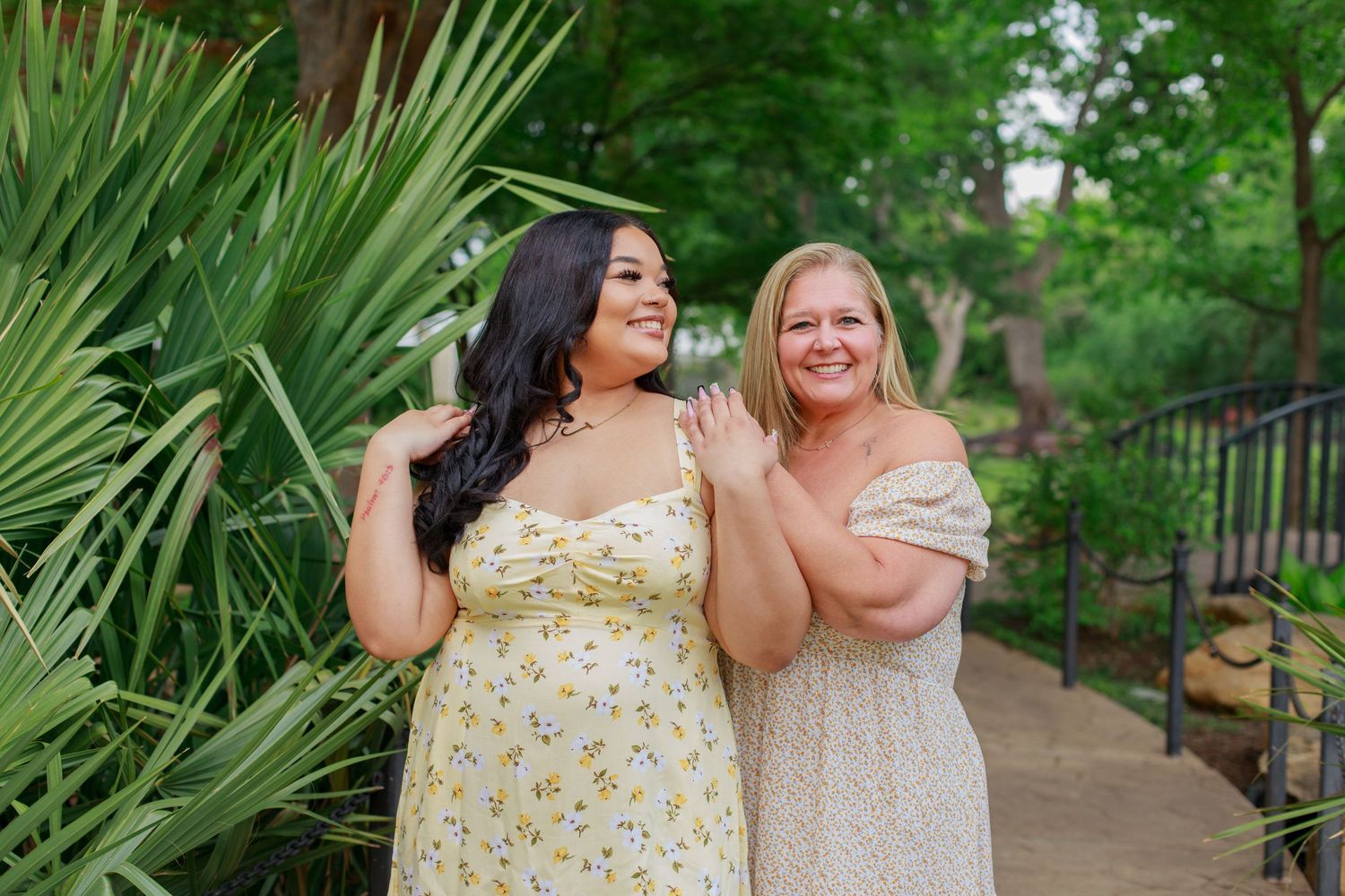 Two women in floral dresses smile and pose together on a garden walkway with palms.