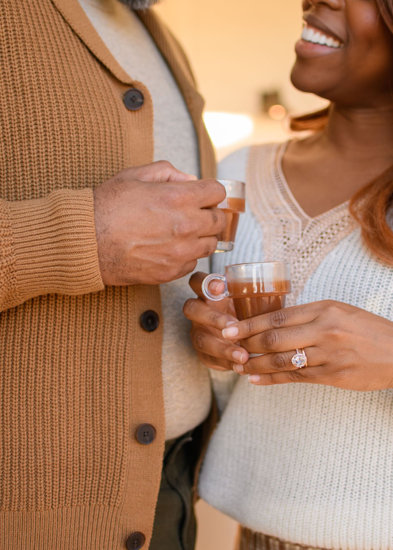 A couple shares a joyful moment holding small drinks, the woman displaying an engagement ring.