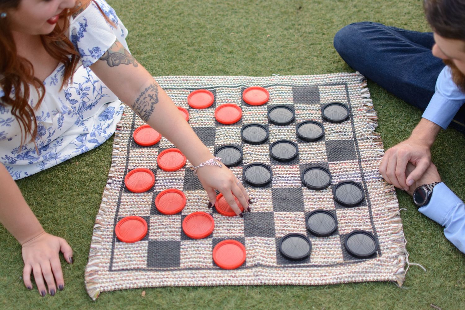 Two people playing a large outdoor checkers game on a woven mat placed on green grass.