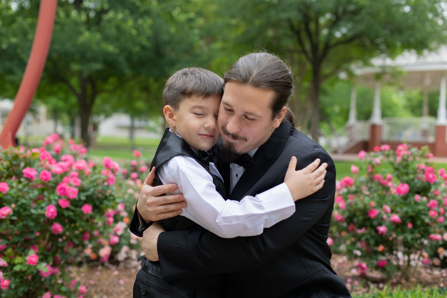 Two people share an emotional embrace outdoors among pink rose bushes.
