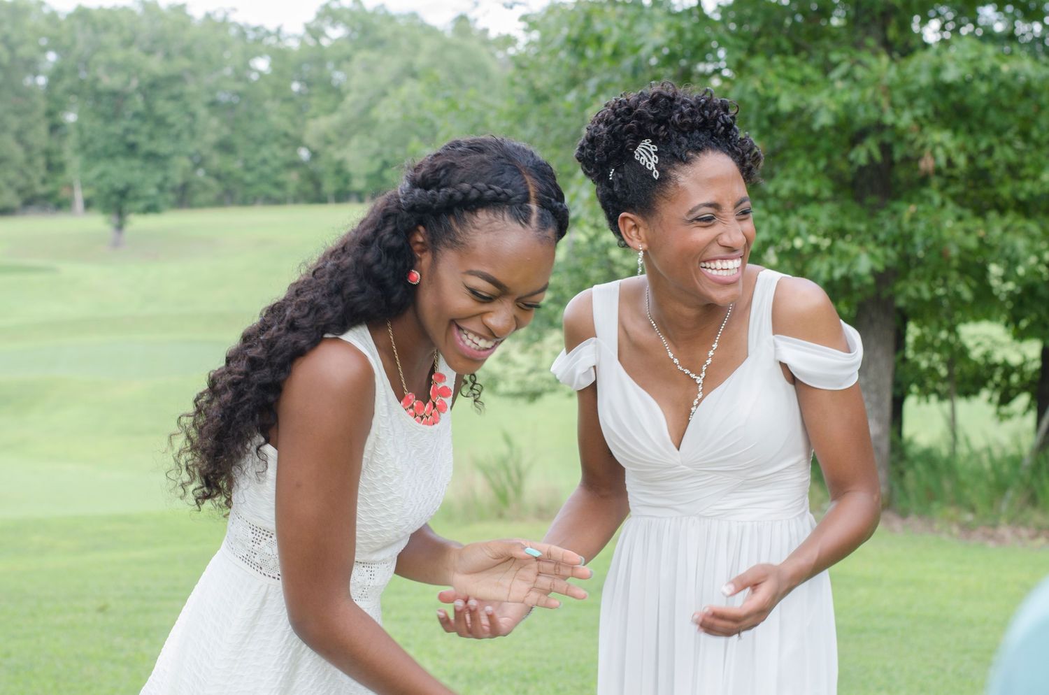 Two bridesmaids in white dresses laughing joyfully together at an outdoor wedding celebration.