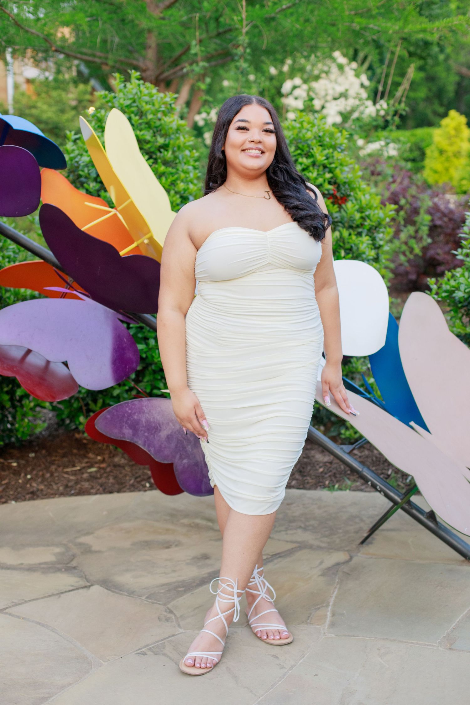 Smiling plus-size woman in white strapless ruched midi dress posing by colorful butterfly art.