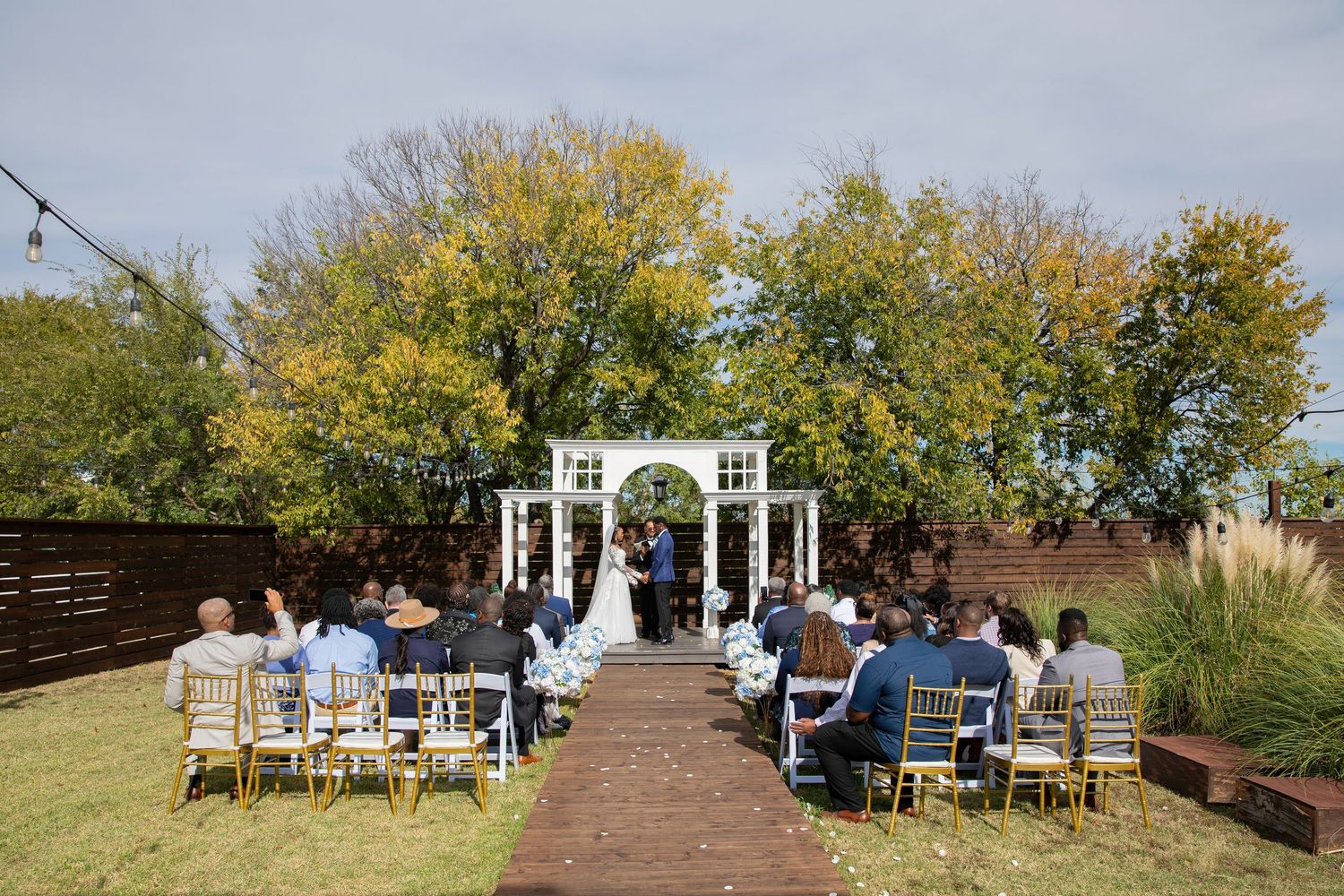 Small outdoor wedding ceremony at a white arbor with guests in gold chairs surrounded by autumn trees.