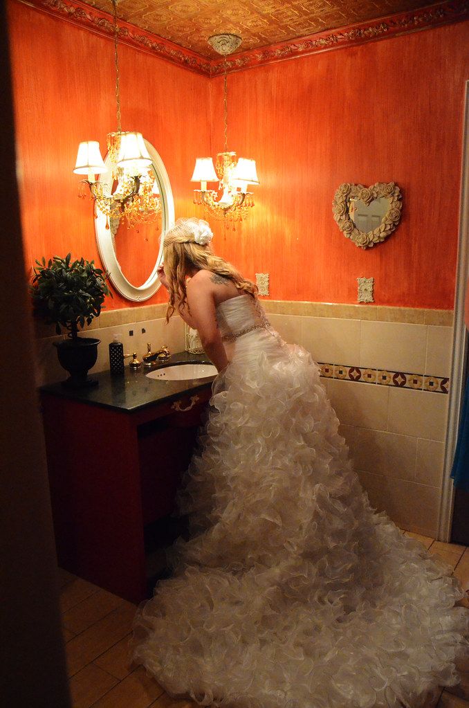 Blonde bride in ruffled white gown looking in oval mirror in ornate orange powder room.