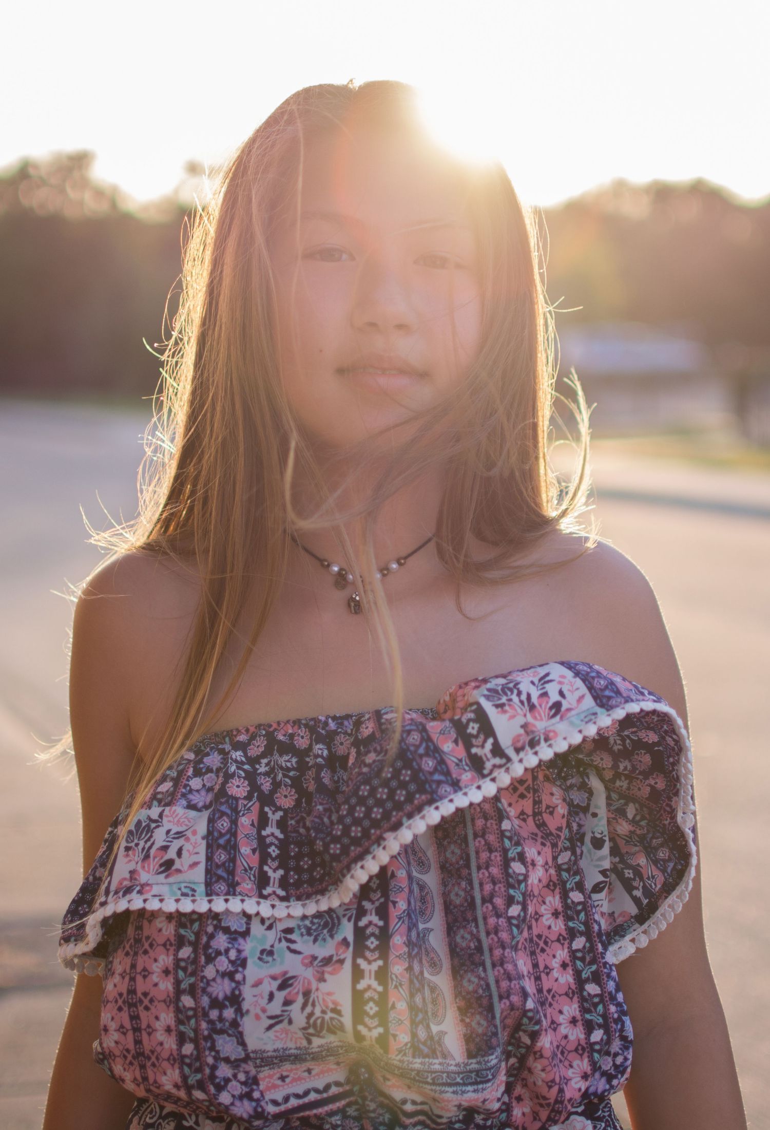 Young girl in floral off-shoulder top stands outdoors with golden sunset backlight behind her.
