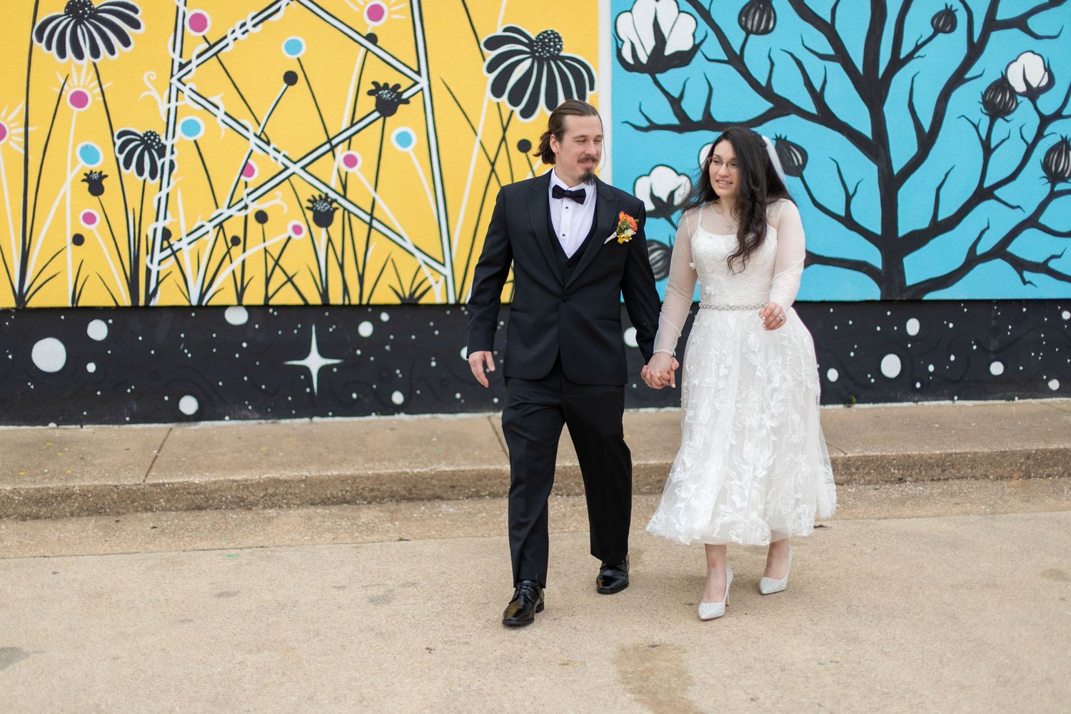 A couple walks together along a sidewalk against a colorful cotton plant mural backdrop.