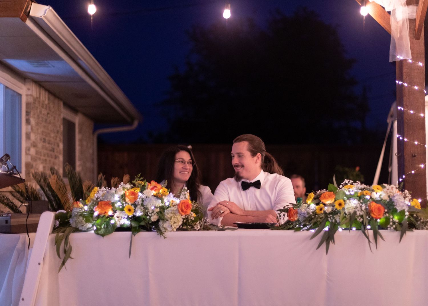 A couple seated at a decorated head table with floral arrangements at night.