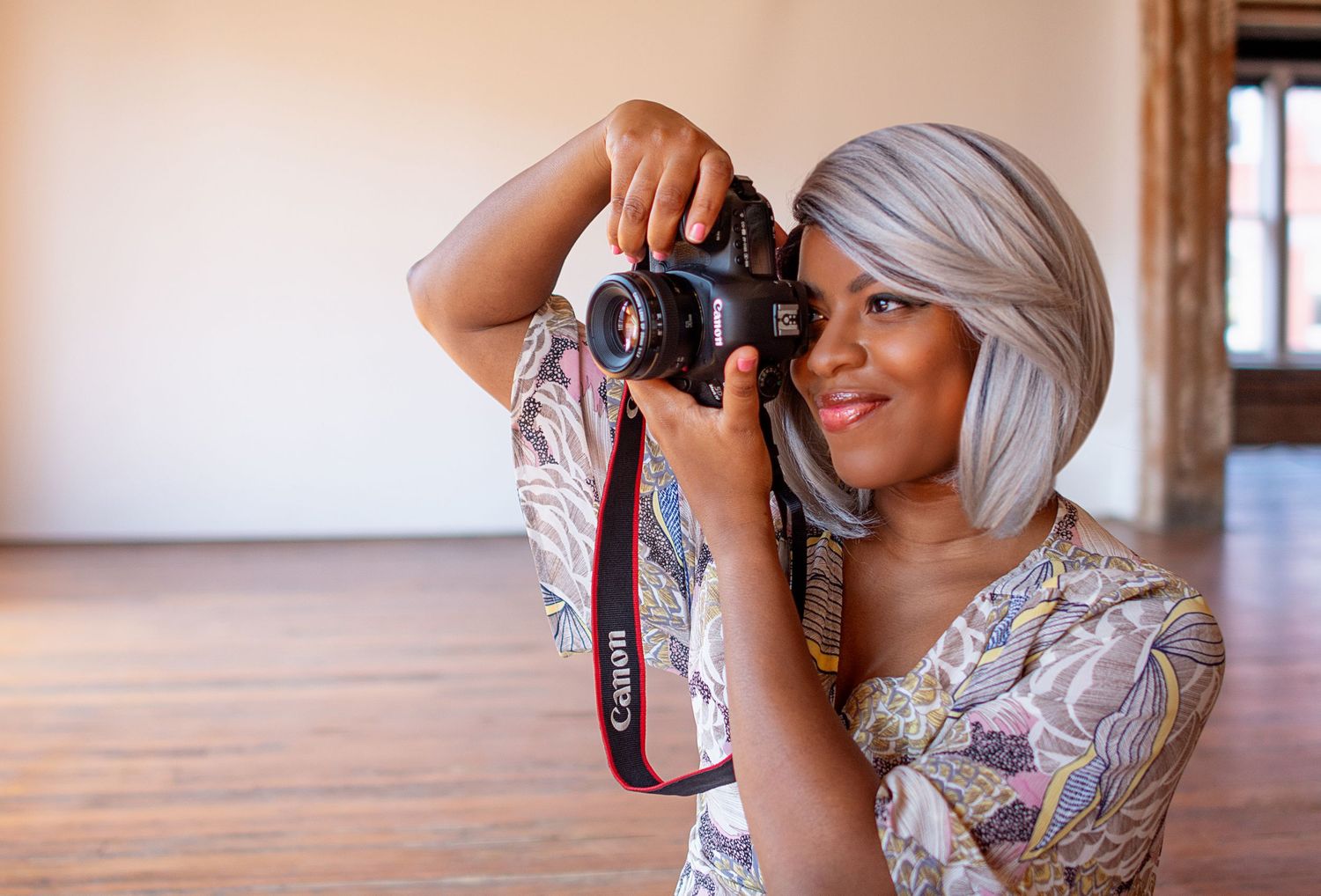 Woman with gray bob haircut holding Canon DSLR camera, wearing floral dress in bright studio space.