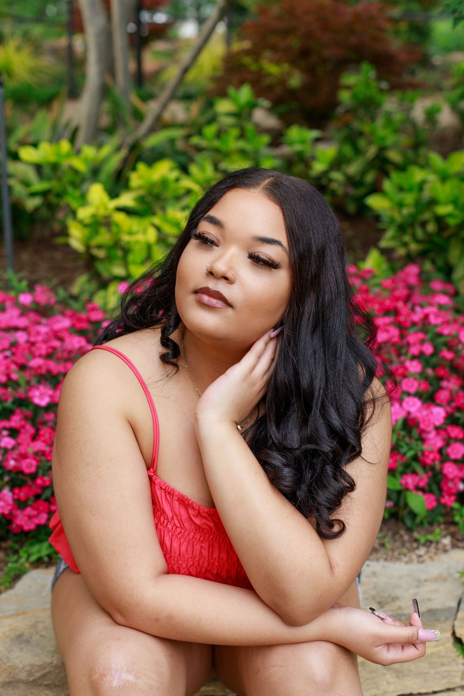 A young woman in a red top sits among pink flowers in a vibrant outdoor garden.