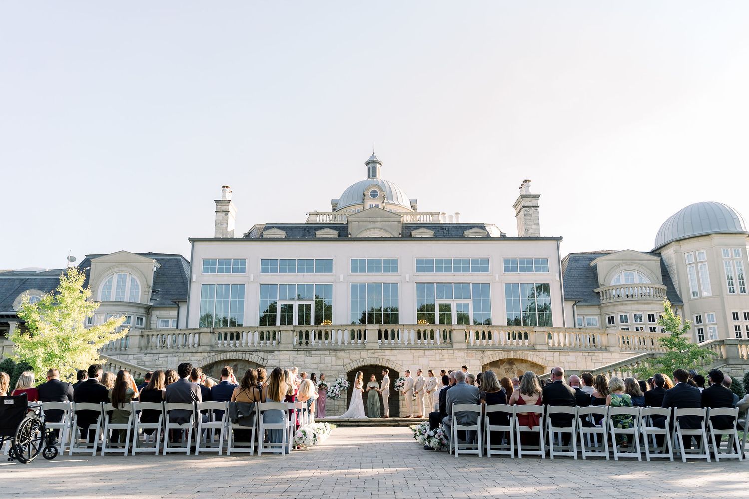 Outdoor wedding ceremony with guests seated in white chairs facing a grand glass-facade estate building on a sunny day.