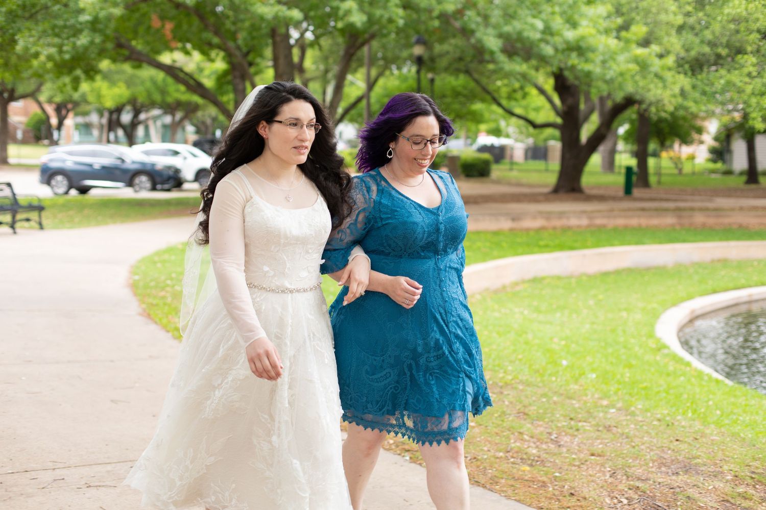 Two people walk arm in arm across a white bridge in a park setting with green trees.