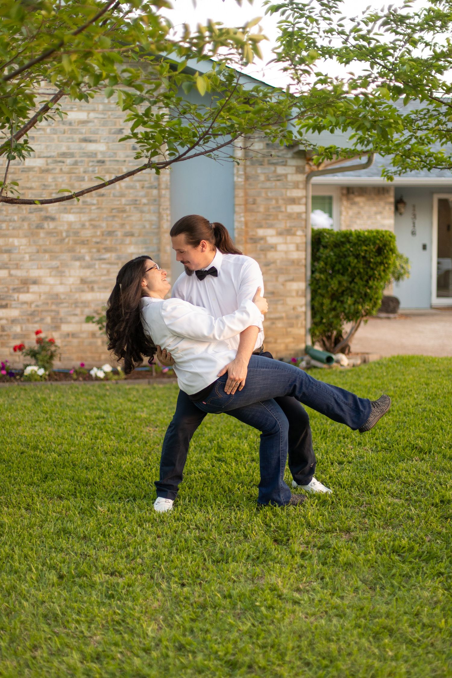 Romantic dip pose on green lawn with brick house and tree in background.