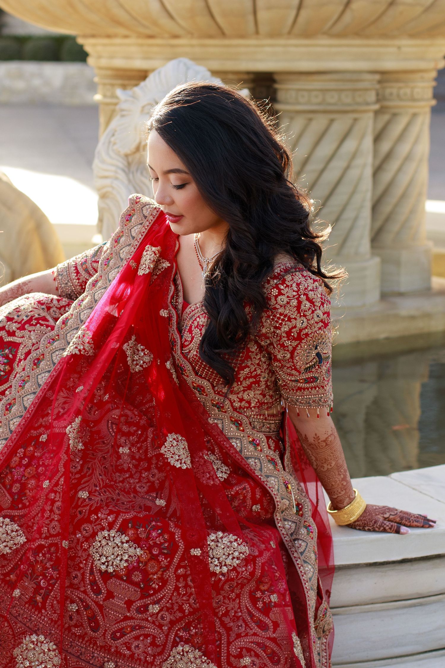 Indian bride in red and gold lehenga with henna hands sits gracefully near an ornate fountain.