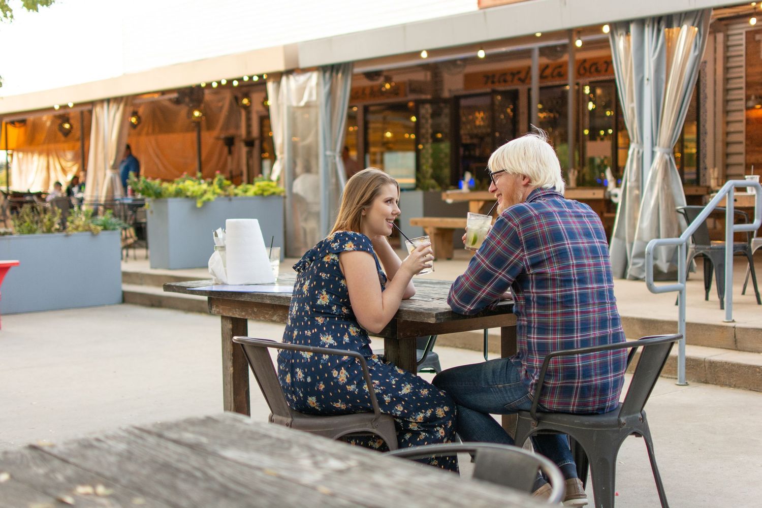 Couple enjoying drinks at an outdoor restaurant patio with string lights at dusk.